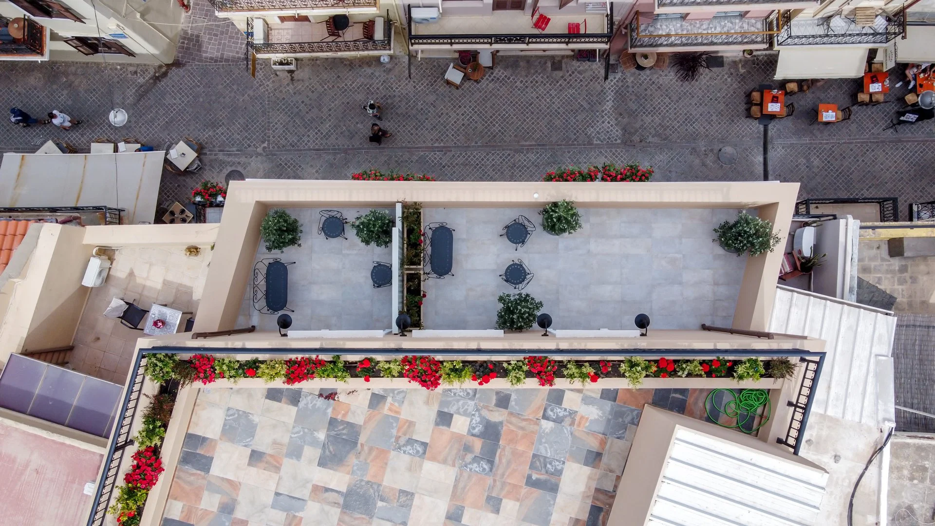 Aerial view of a rooftop patio with outdoor furniture, potted plants, flowers, and a glass wall, overlooking a cobblestone street with pedestrians and outdoor dining tables.