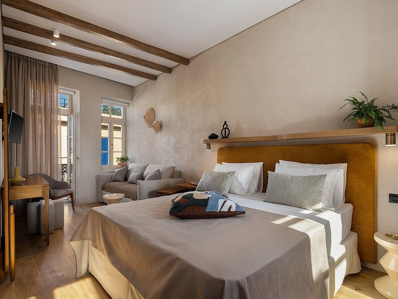 A bedroom with a large bed, two pillows, and a decorative pillow, a wall shelf with decor, a window with natural light, a sitting area with a sofa and armchair, a desk with a chair, and plants.