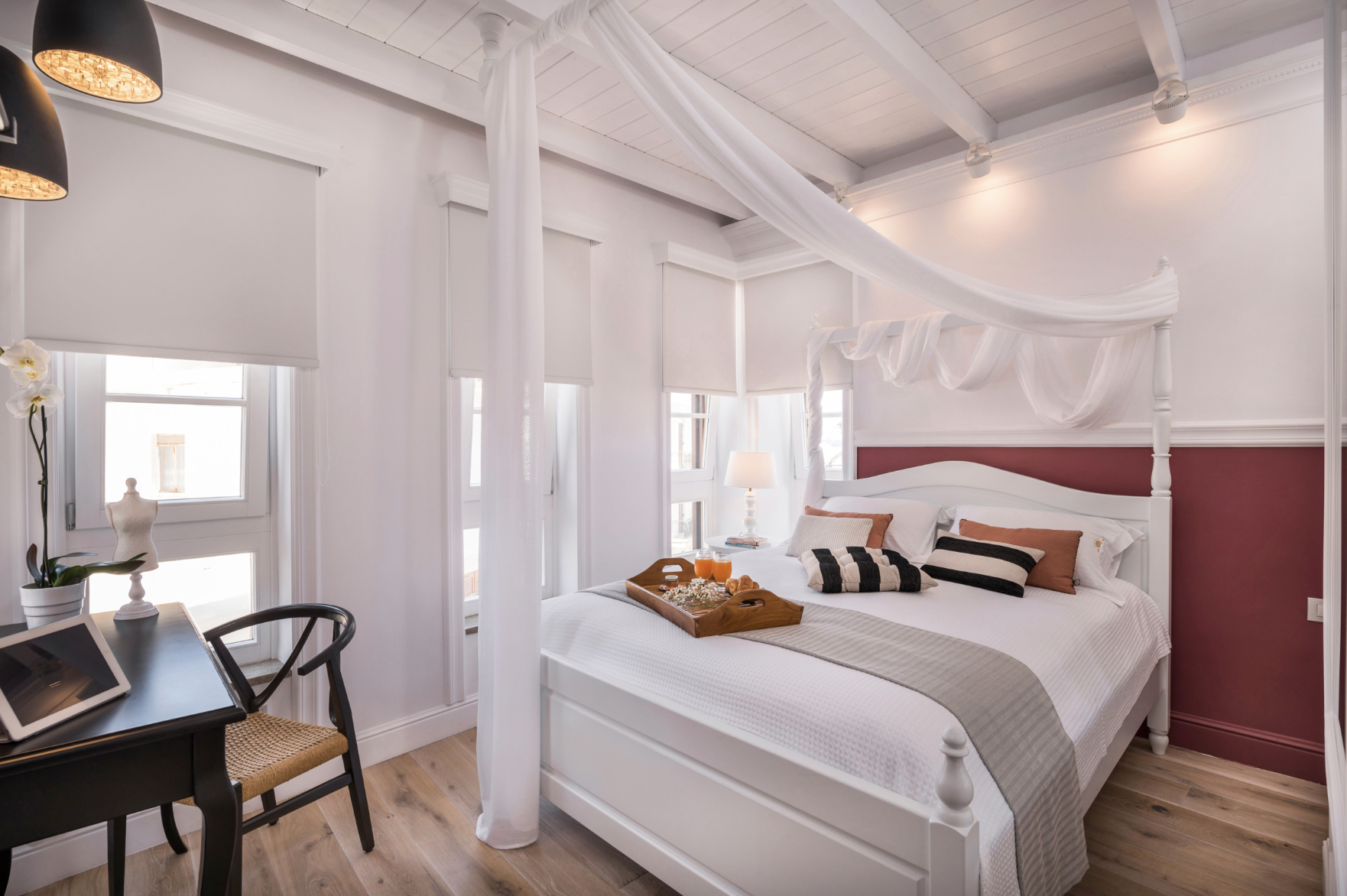 A bedroom with a white four-poster bed, decorated with pillows, a tray with breakfast, and a sheer canopy. The room has white walls, several windows with white blinds, a white ceiling with exposed beams, and wooden floors. There is a small black table with a tablet and a chair, a side table with a lamp, and a mannequin on the windowsill.