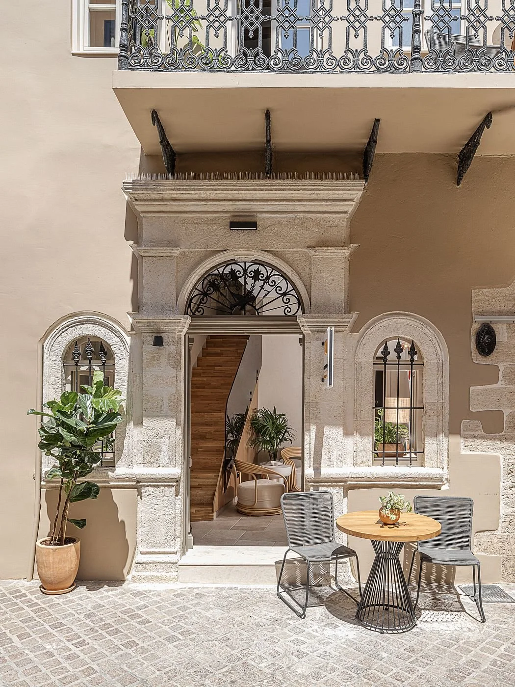 Entrance to a boutique hotel with arched windows and stonework, potted plants, patio table and chairs, and a staircase inside.