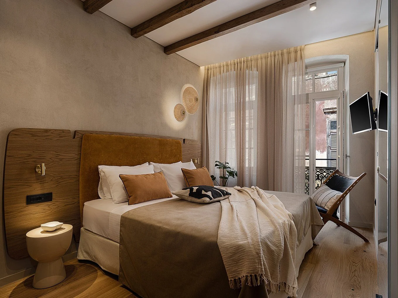 A cozy bedroom with a bed featuring brown and white pillows, a beige throw blanket, and a fabric headboard. The room has wooden ceiling beams, sheer curtains, a balcony door, wall-mounted TV, side table, and a wooden chair.