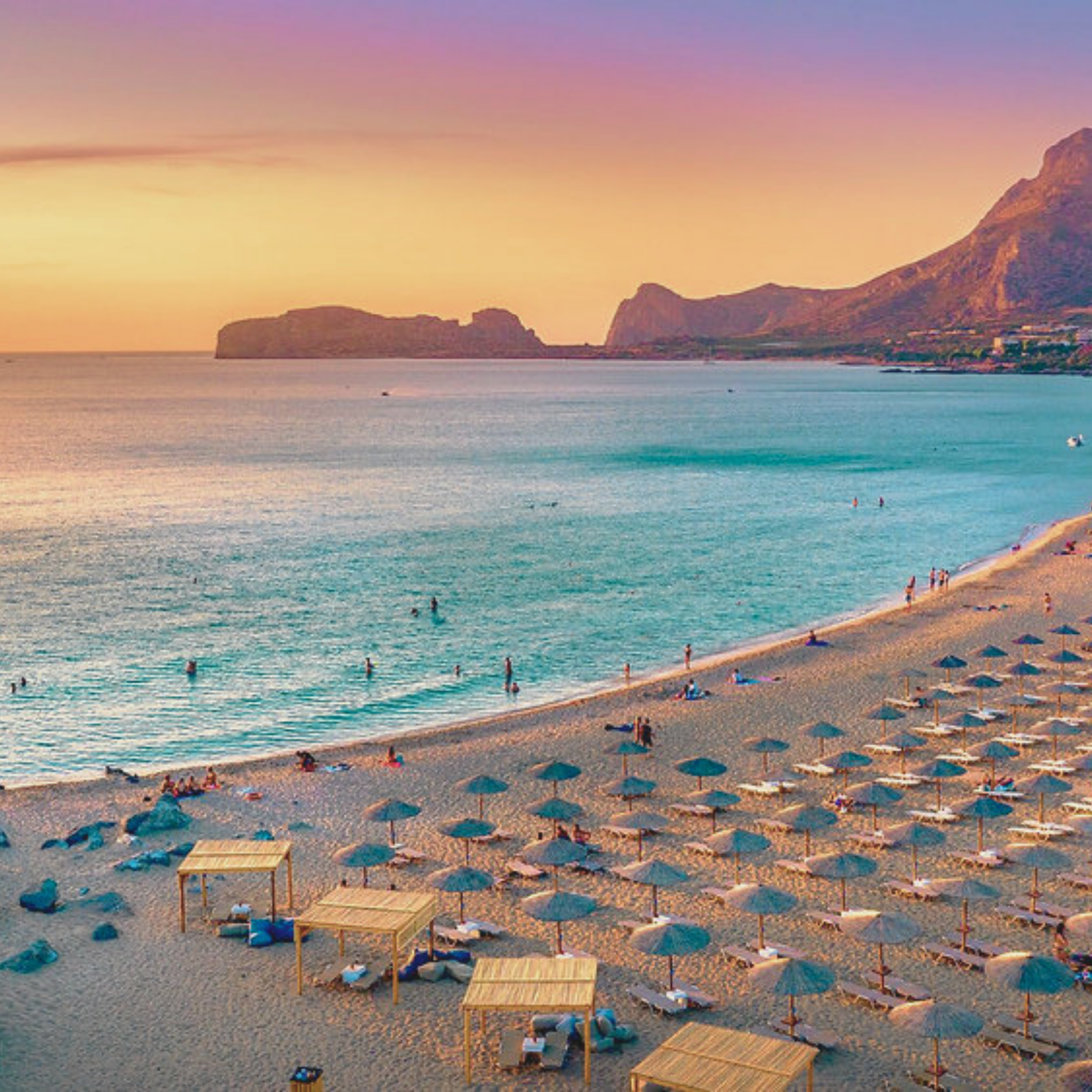 A beach scene during sunset with small wooden cabanas and umbrellas along the shoreline, showing calm water and distant cliffs or mountains.