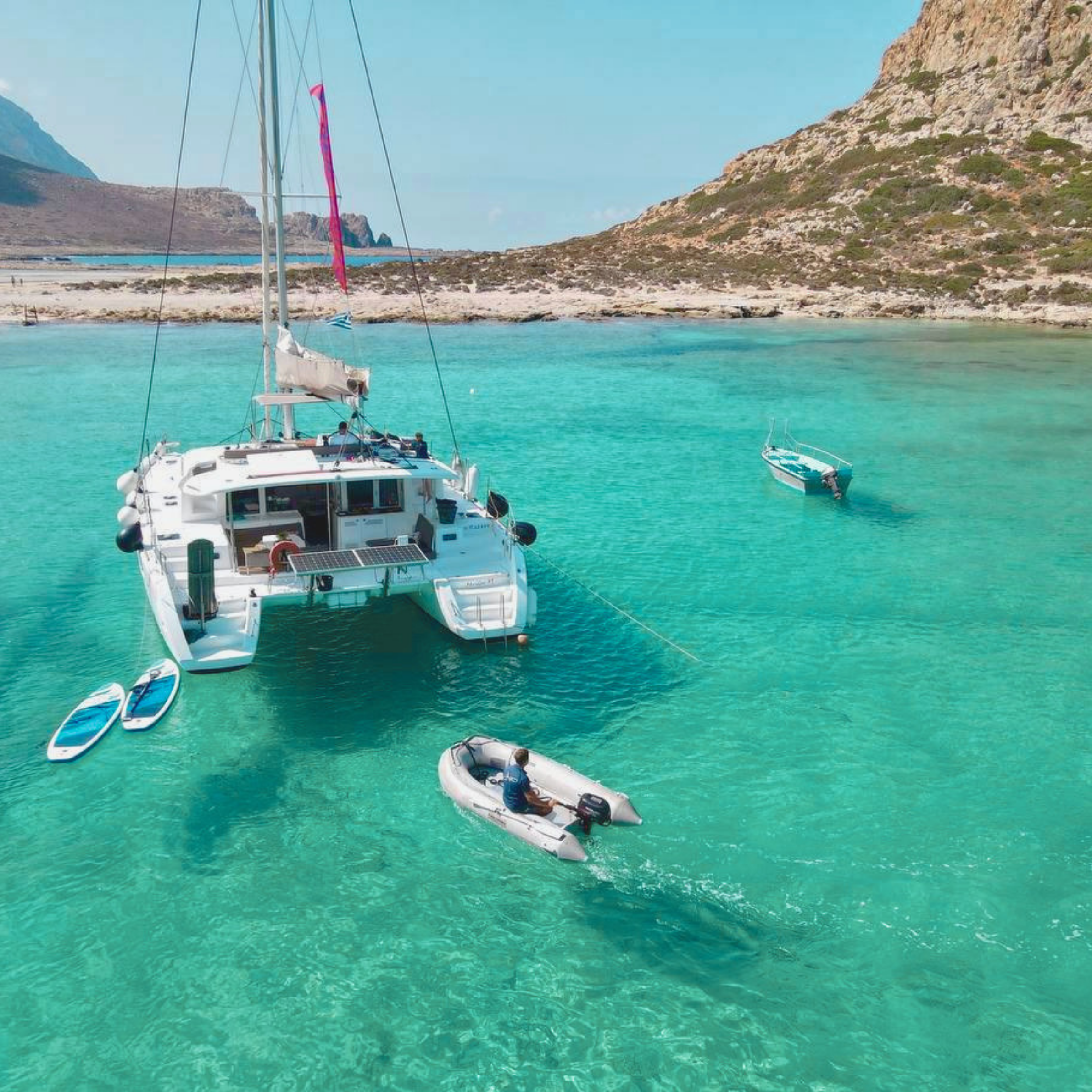 A white sailing yacht and small paddleboards floating on clear turquoise water near a rocky shoreline and mountainous landscape.
