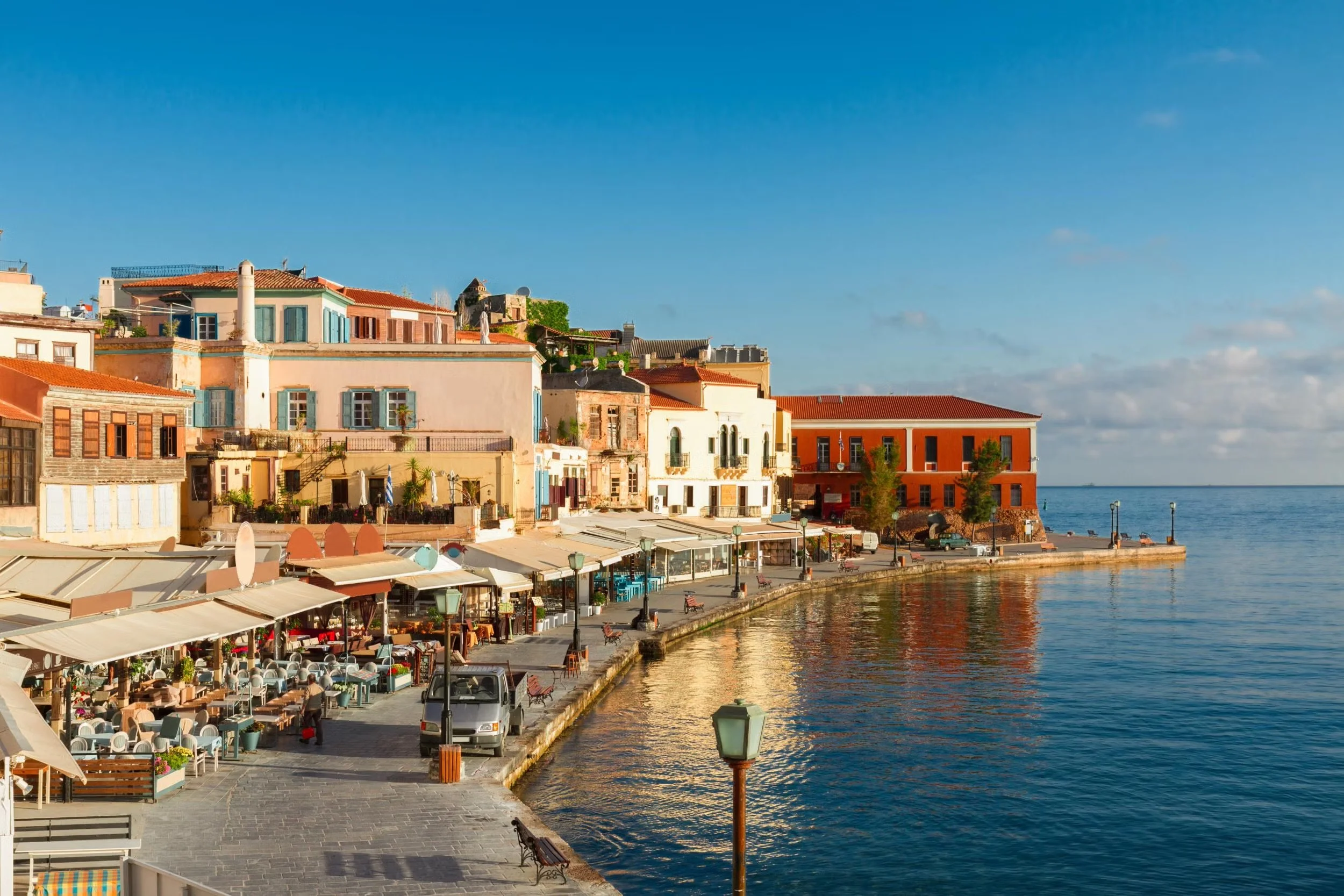 A waterfront promenade with outdoor dining, benches, lamp posts, and a view of buildings and the water under a blue sky.