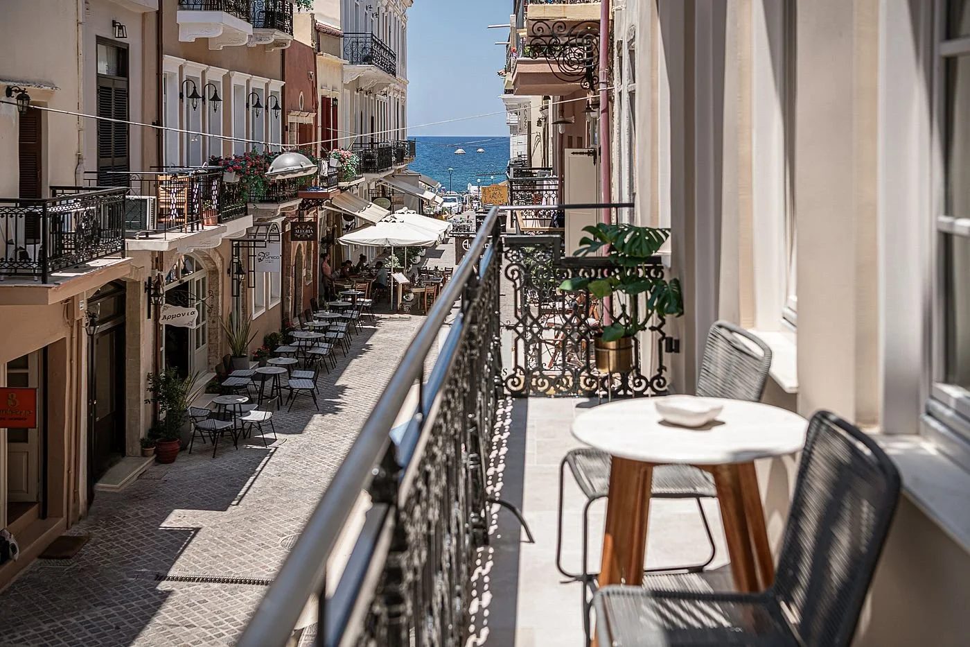 View from a balcony overlooking a narrow street with outdoor cafes and shops, leading to the sea in the background.