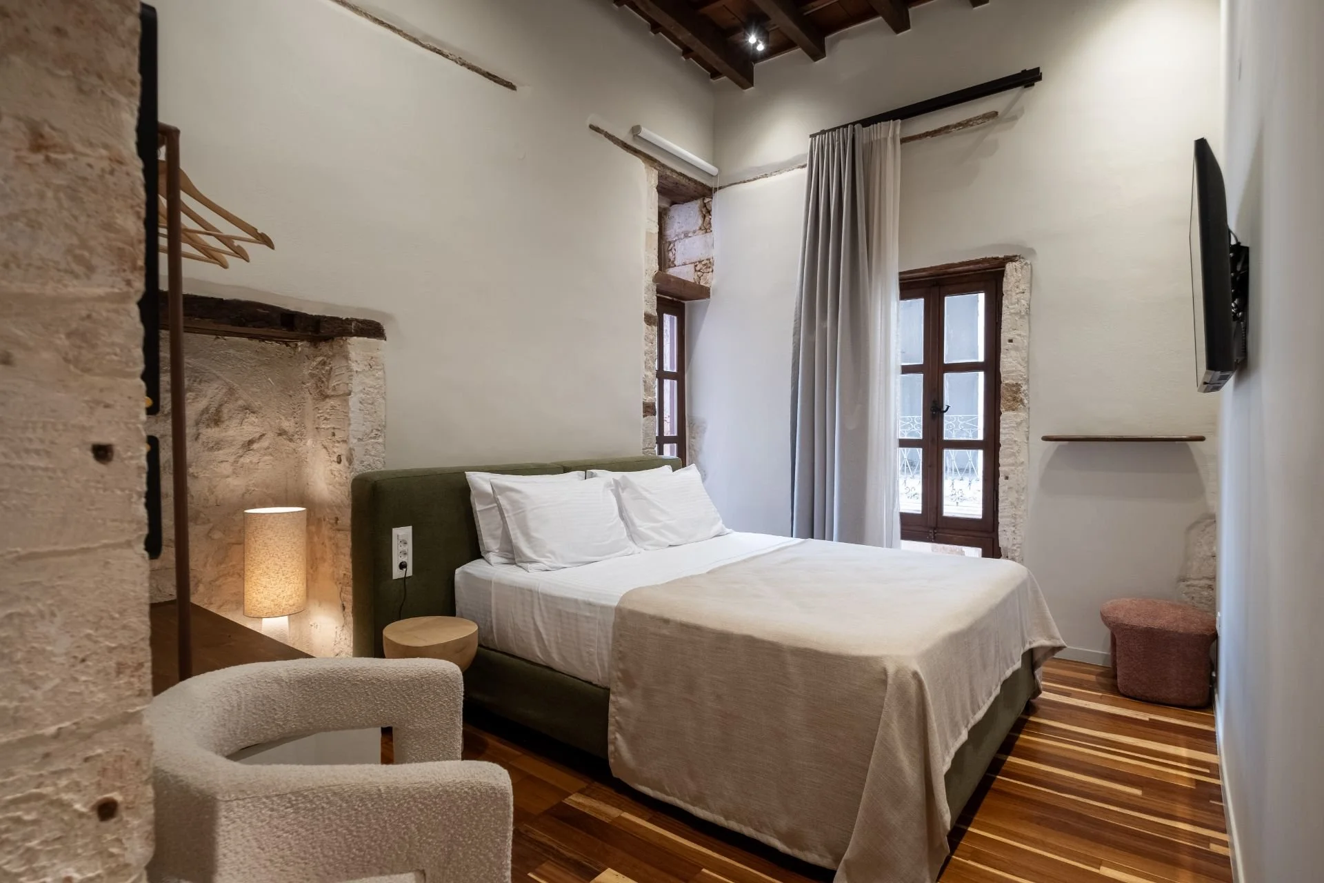 A cozy bedroom with a bed that has a green headboard, white bedding, and pillows. There are two windows with wooden frames, a curtain, and exposed brickwork around the windows. A lamp, a small round table, and an armchair are also visible. The ceiling has wooden beams, and there is a TV mounted on the wall.