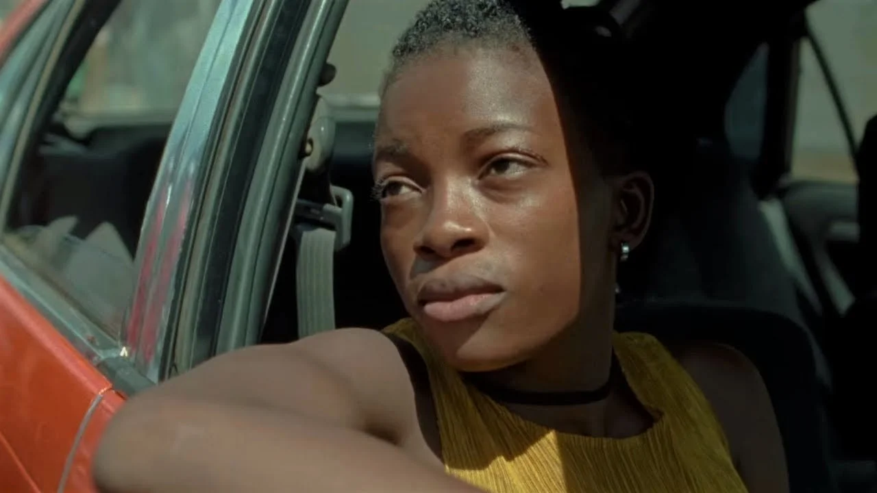 A woman with short hair, earrings, and a yellow top looking out of a car window with her arm resting on the door.