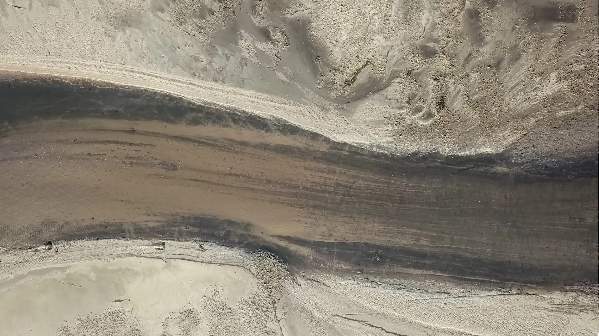 An aerial view of a sandy beach with a winding waterway or wetland area running through the sand, showing different textures and colors of sand and water.