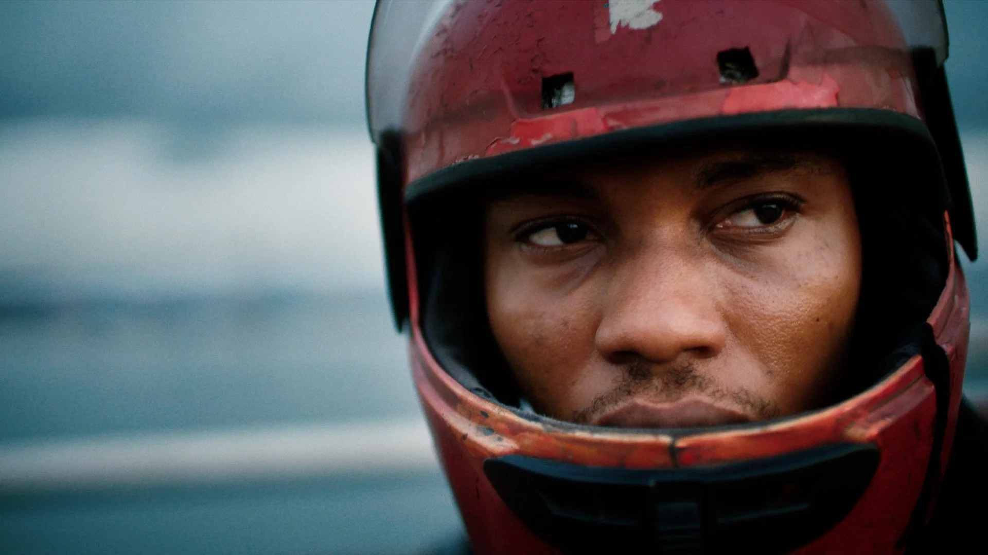 Close-up of a man wearing a red helmet, with a focused expression, standing outdoors with a blurred blue sky and ocean in the background.