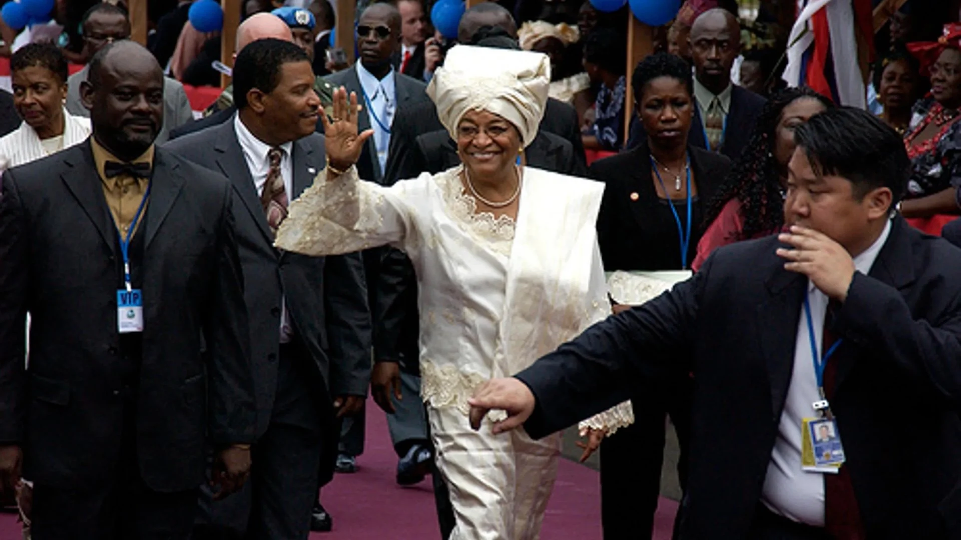 A woman dressed in traditional attire and a headwrap, smiling and waving at a formal event, surrounded by other people in suits and dresses, some wearing lanyards and VIP badges.