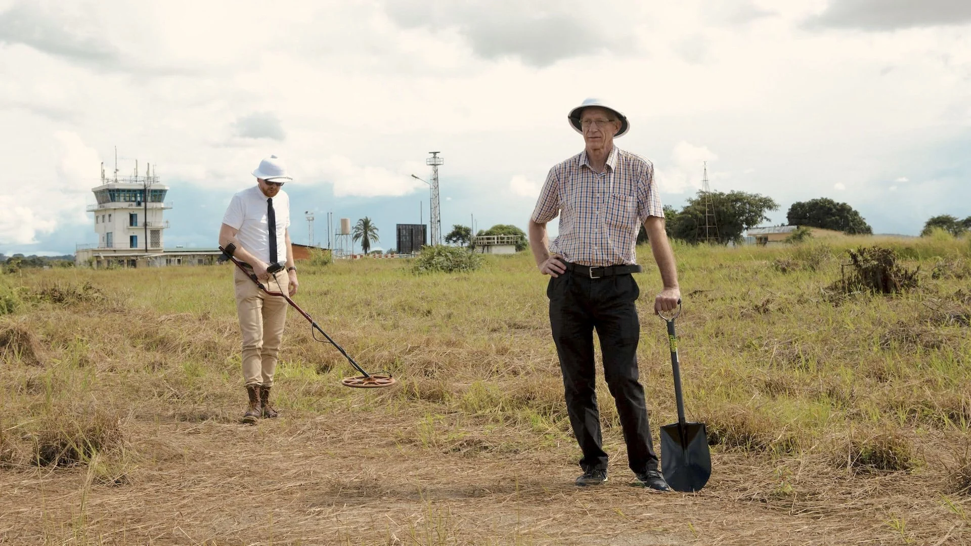 Two men in hats standing in an open grassy field, with one holding a metal detector and the other a shovel, under a cloudy sky.