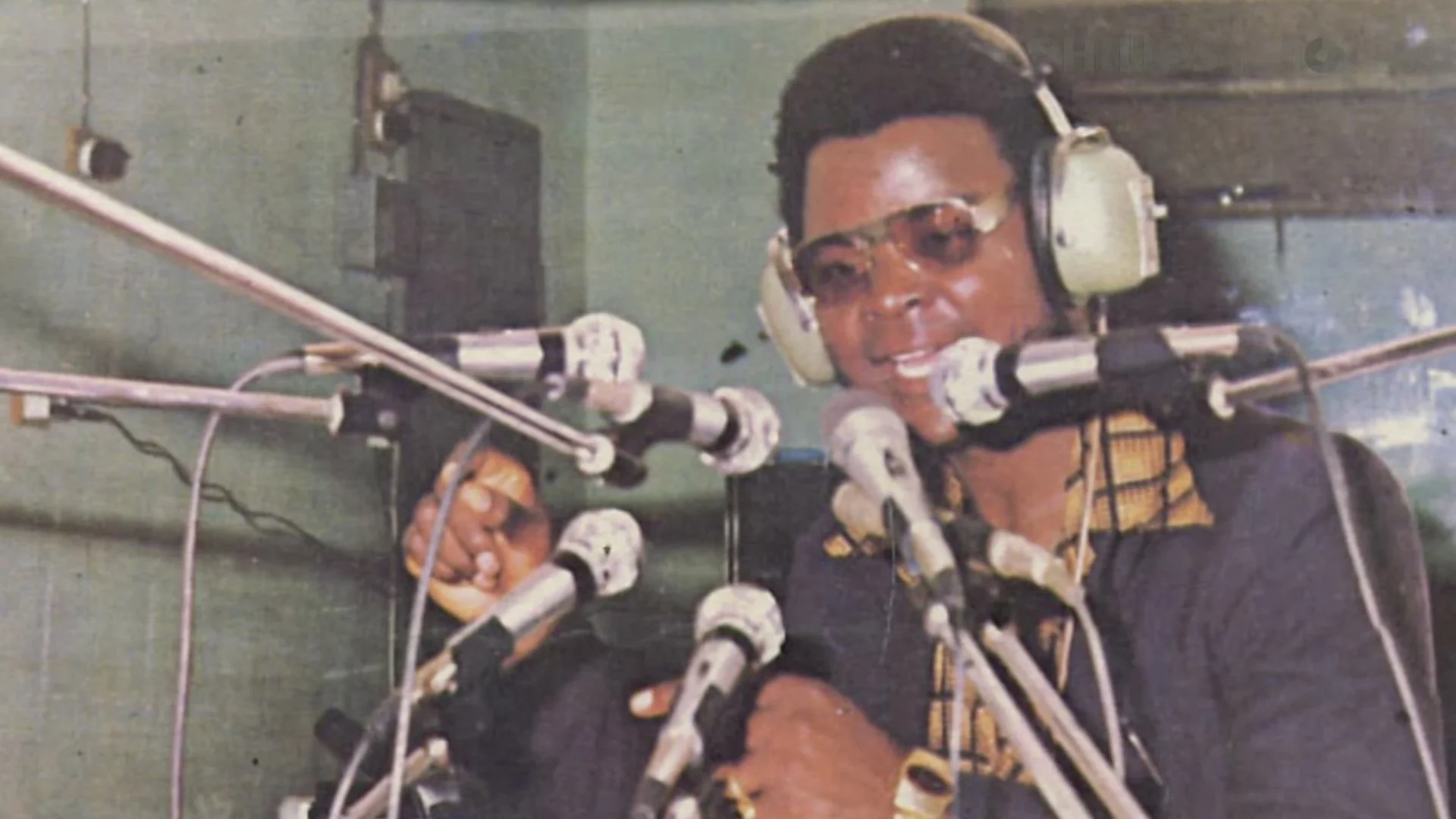 A man wearing headphones and sunglasses speaking into multiple microphones in a recording studio.