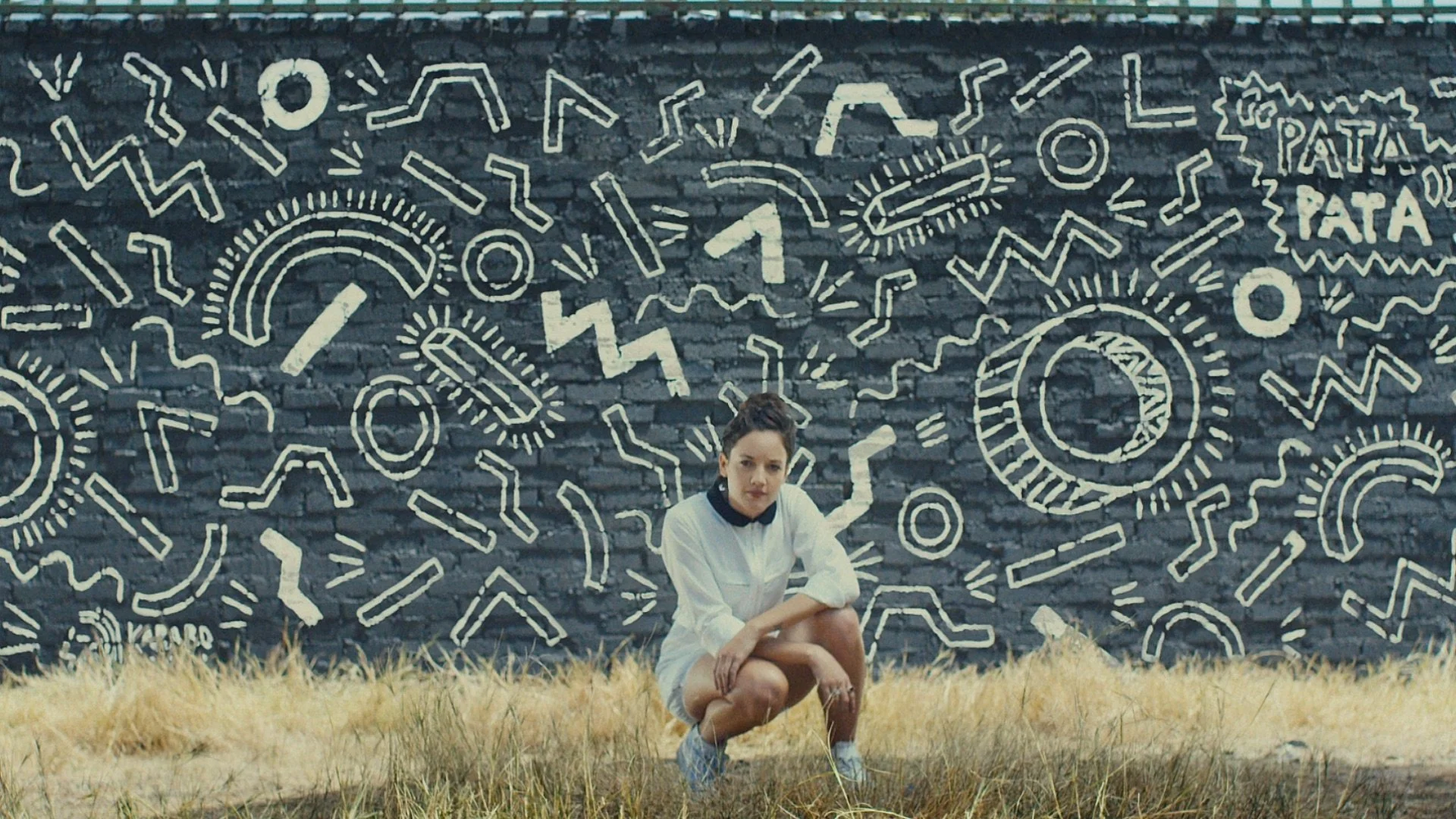 A woman squats in front of a large black brick wall decorated with white abstract line drawings, including rainbows, zigzags, circles, and the text 'Patatap Patan.'