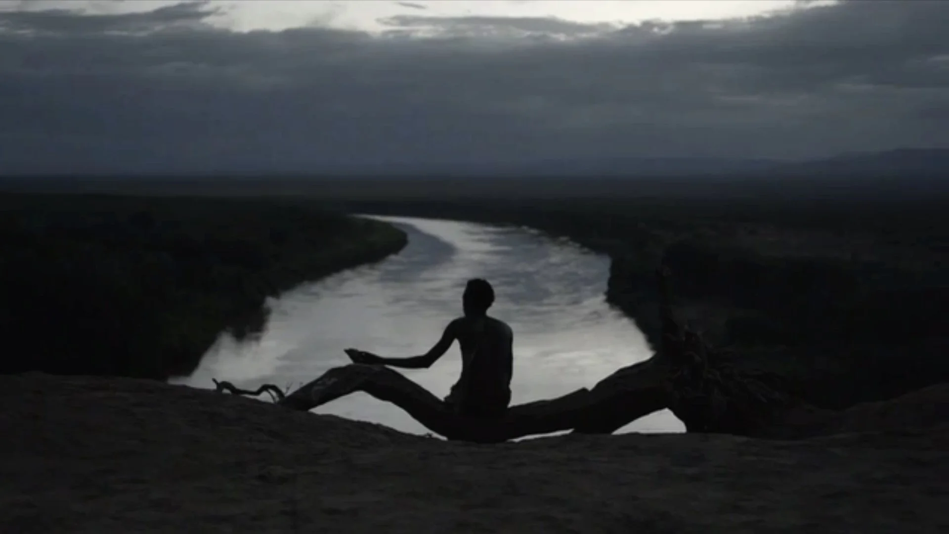 Silhouette of a person sitting on a fallen tree branch by a river at dusk, with dark clouds overhead.