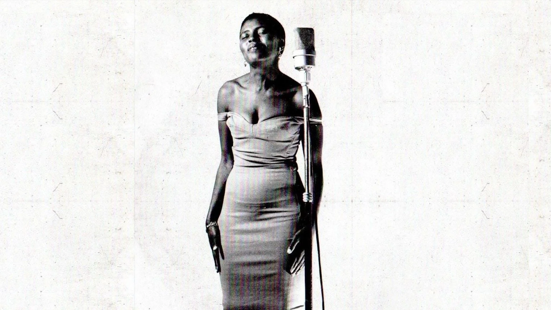 Black and white photo of a woman singing into a vintage microphone, wearing an elegant off-shoulder gown, with her eyes closed, in front of a plain white background.