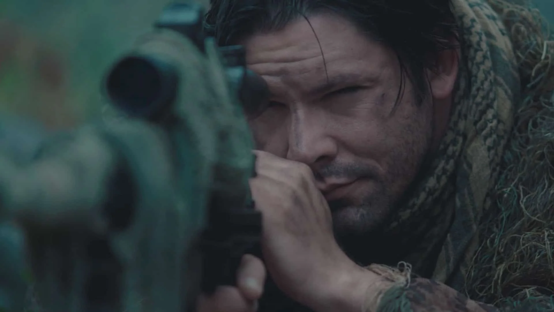 A soldier lying on the ground, aiming a rifle through its sights, wearing camouflage clothing, with a focused expression.