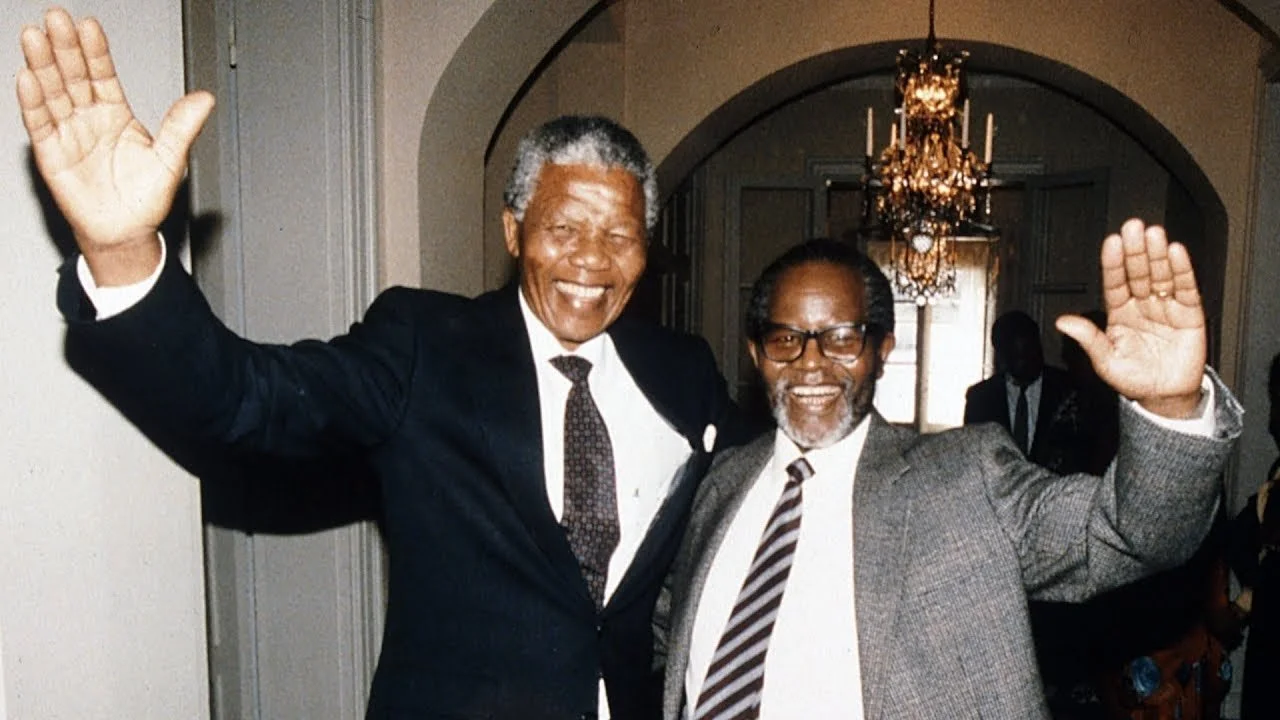 Two smiling men in suits waving, standing indoors at a formal event with a chandelier and guests in the background.