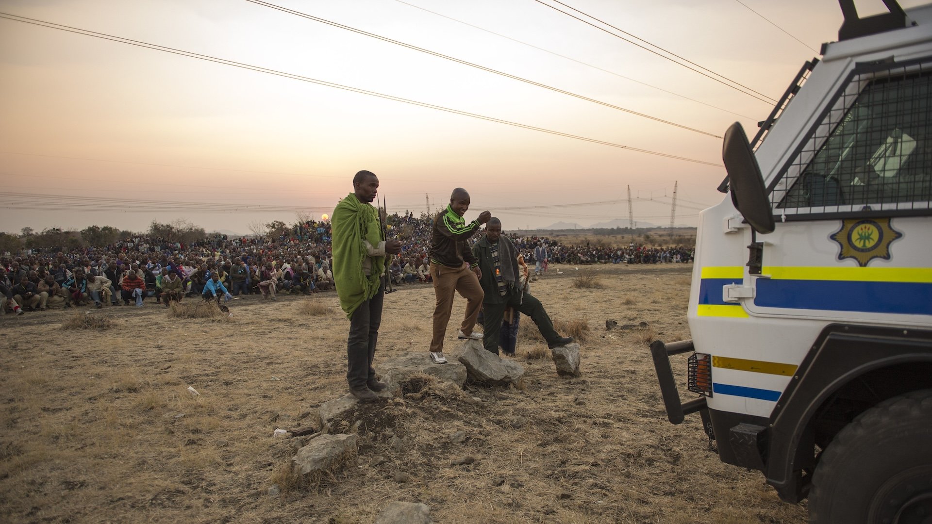 Large crowd of people gathered outdoors during sunset, with three men standing on rocks near a police vehicle.