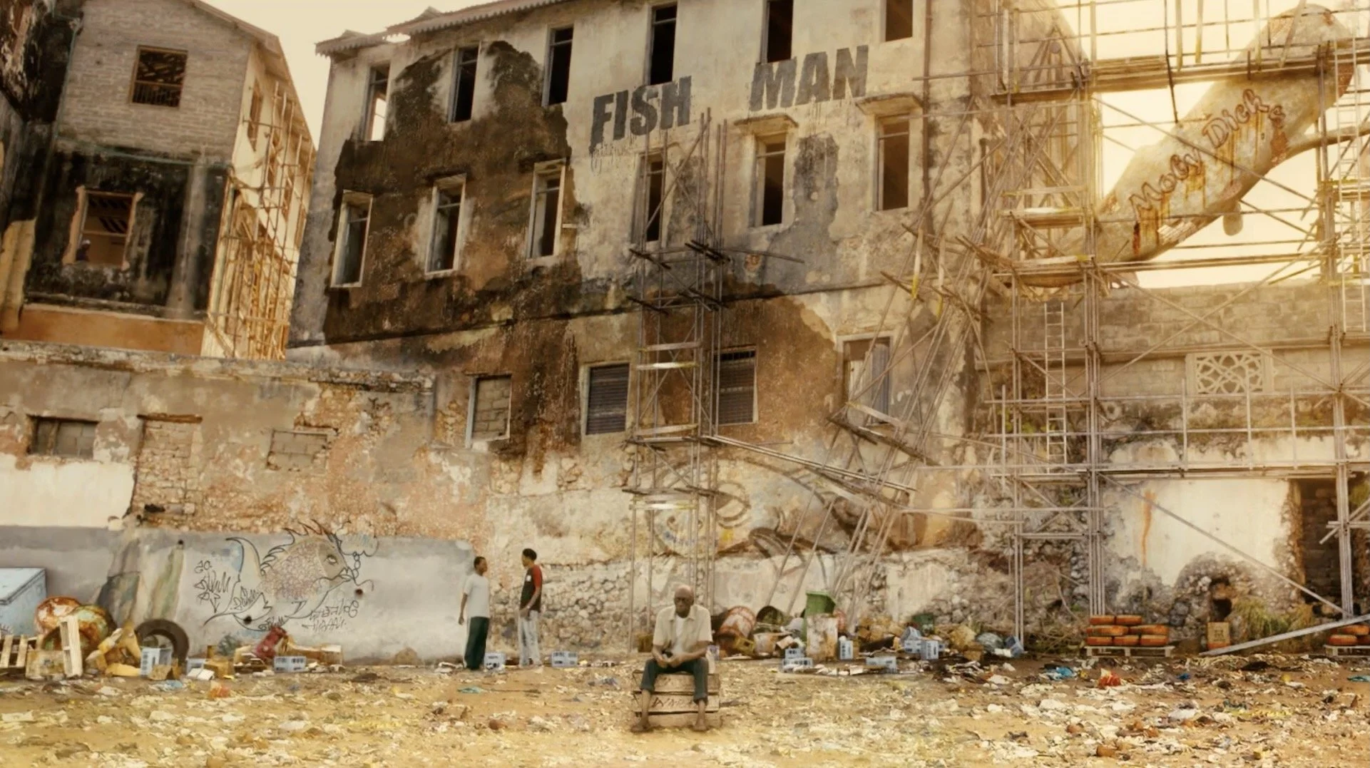 Man sitting on a crate in front of an abandoned, graffiti-covered building with broken windows, scaffolding, and debris on the ground.
