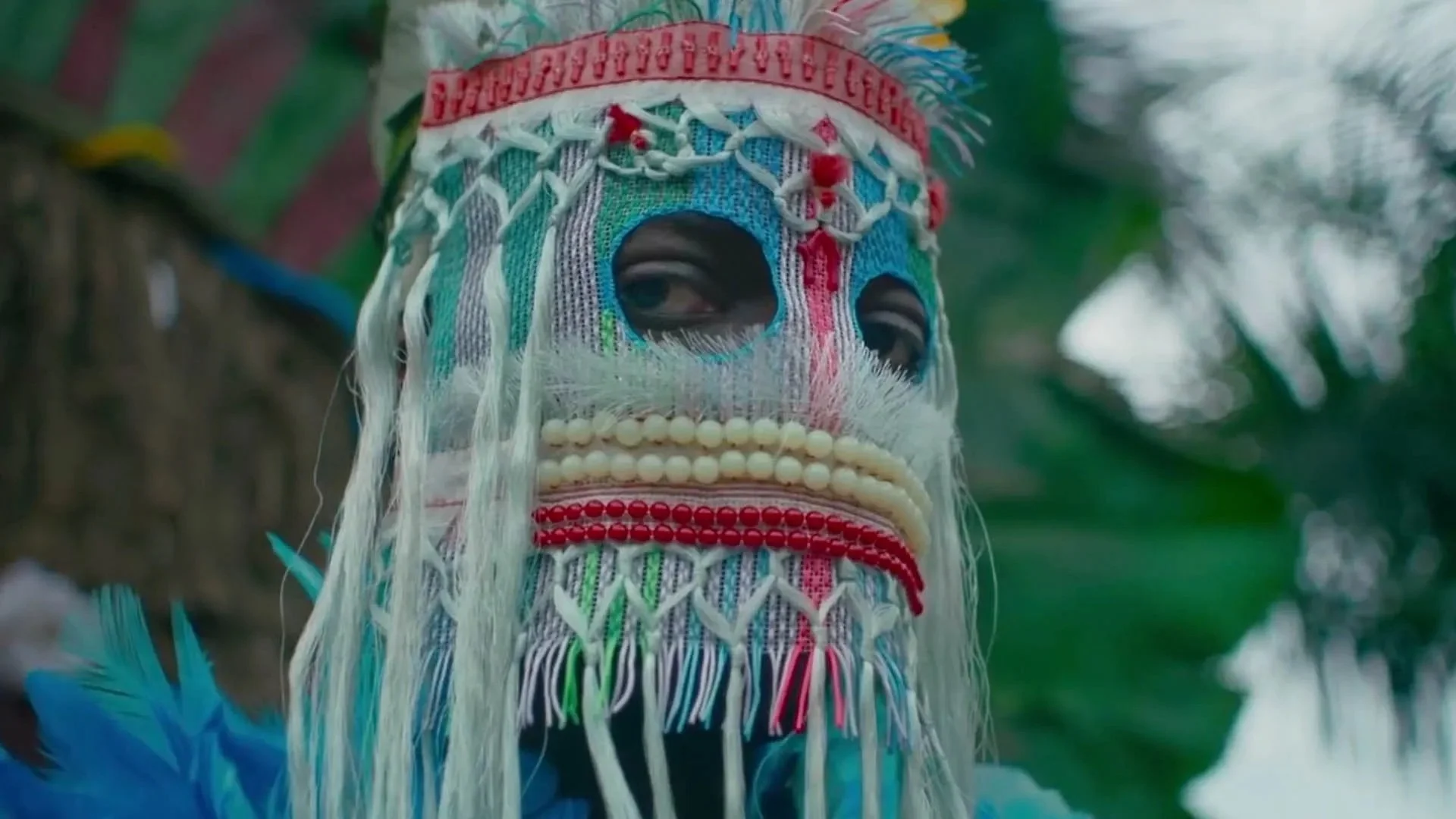 A person wearing a colorful, traditional mask with intricate embroidery, beads, and feathers, outdoors with green foliage.