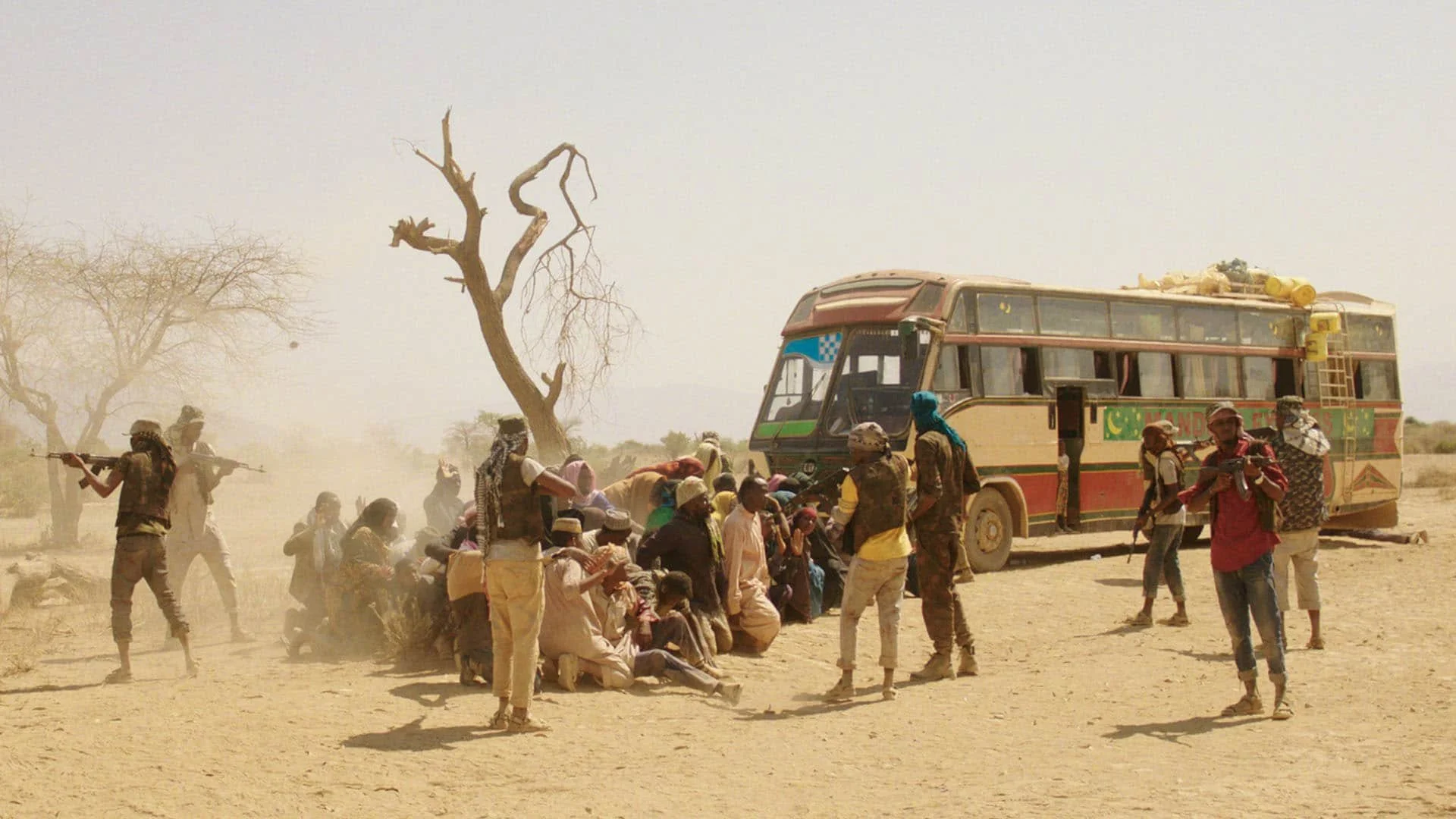A group of people gather around a colorful bus in a dry, dusty landscape with leafless trees, some carrying supplies, some standing, some sitting, with armed individuals present.