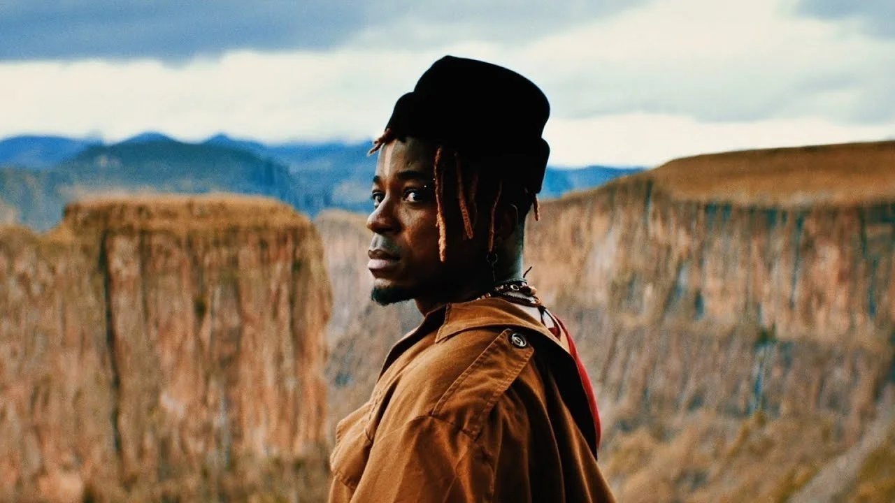 A man with dreadlocks wearing a black hat and brown jacket standing in front of a canyon landscape with mountains in the background.