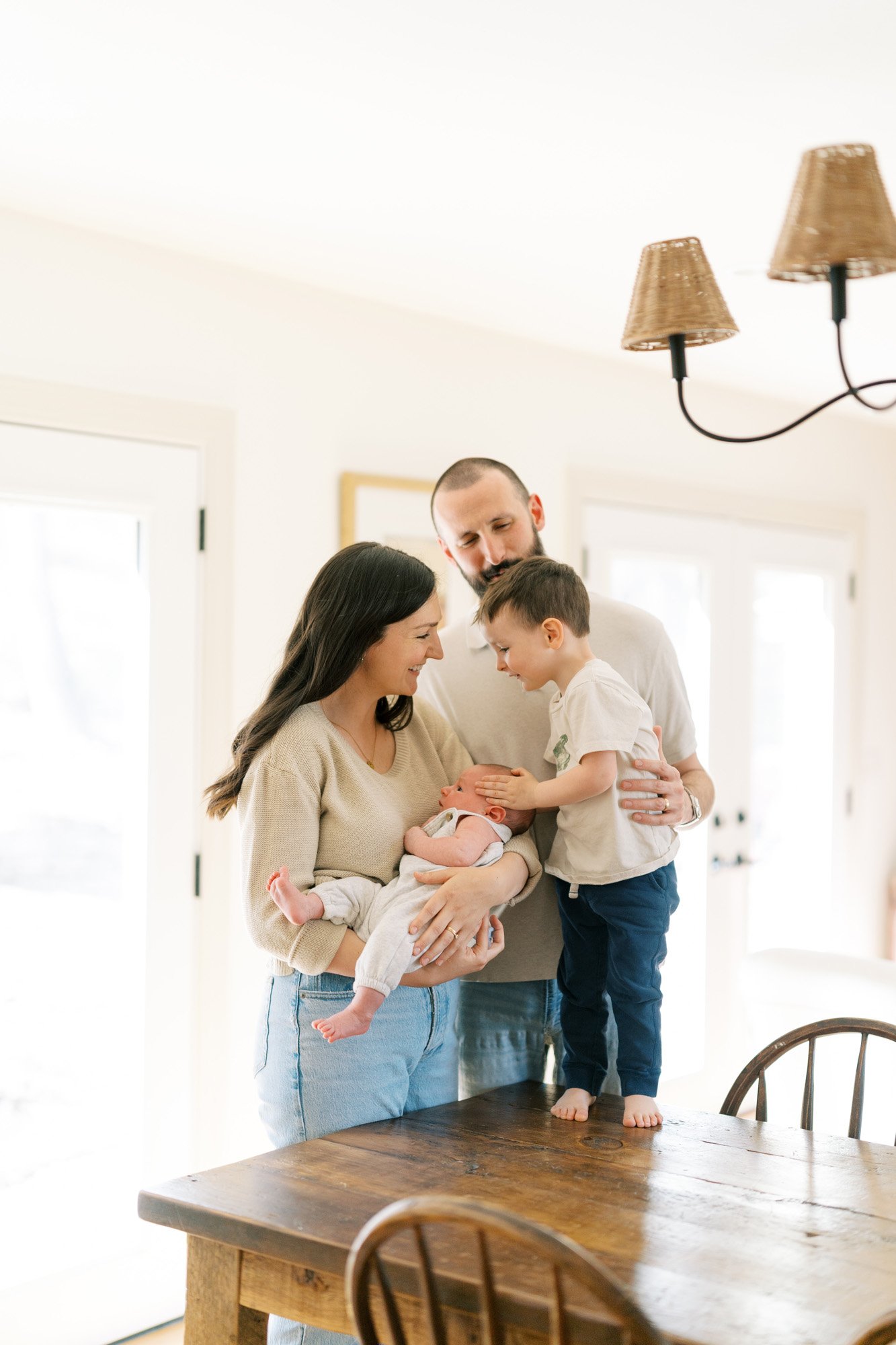Mom holding baby and dad standing near kitchen table, preschool brother standing on table with hand on sibling's head