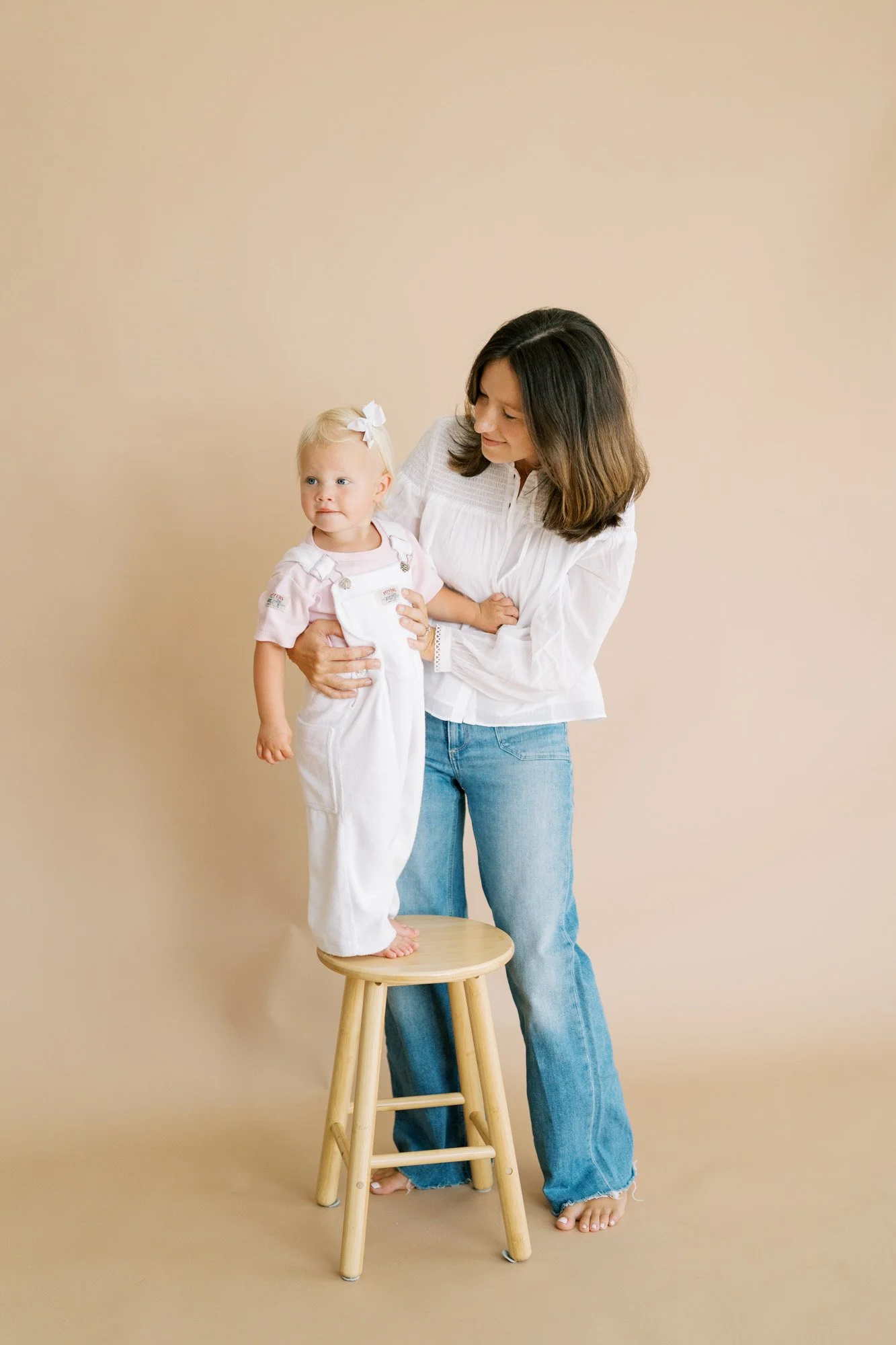 Mom holding infant daughter standing on brown stool in front of a paper backdrop in studio