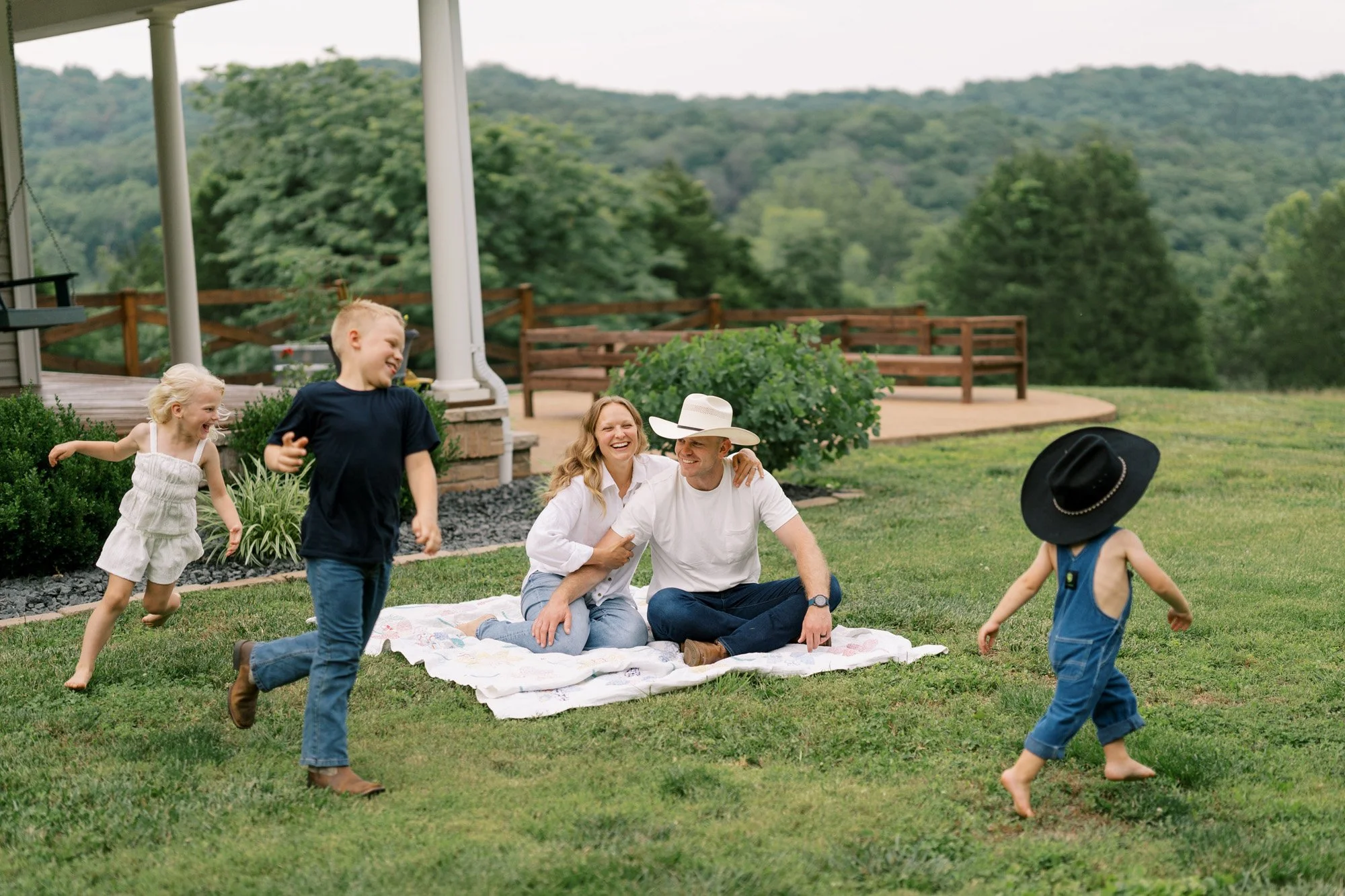 Parents sitting on blanket in their yard while three kids run happily around