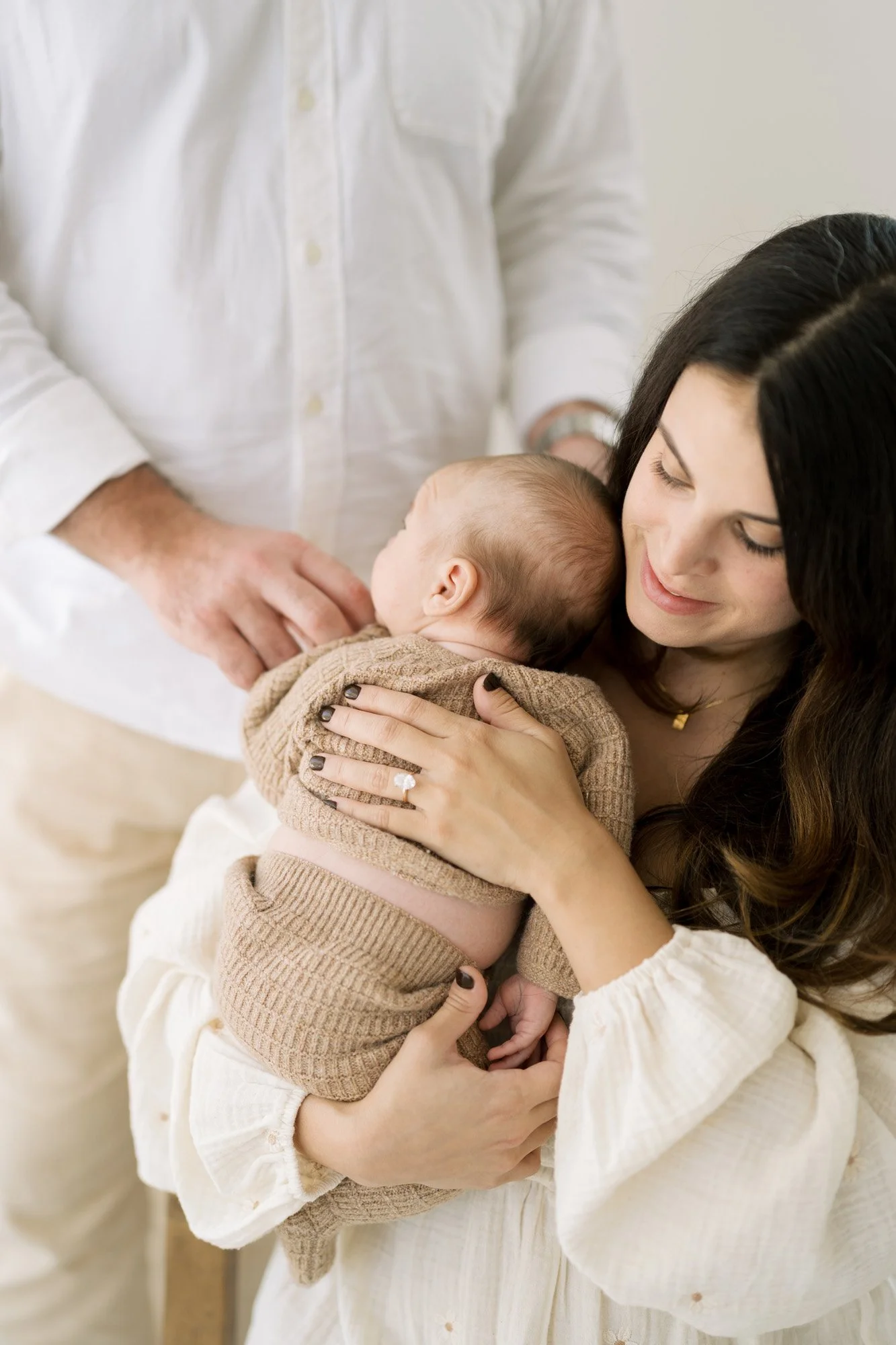 Dad standing behind mom holding newborn near window in studio