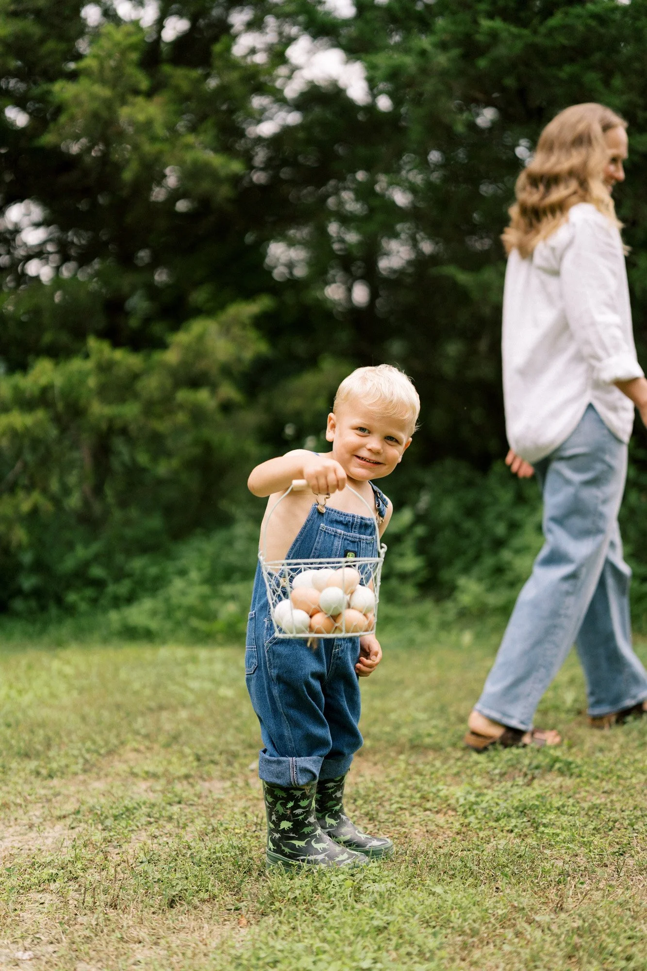 Boy in overalls and boots holding metal basket of eggs with mom in the background