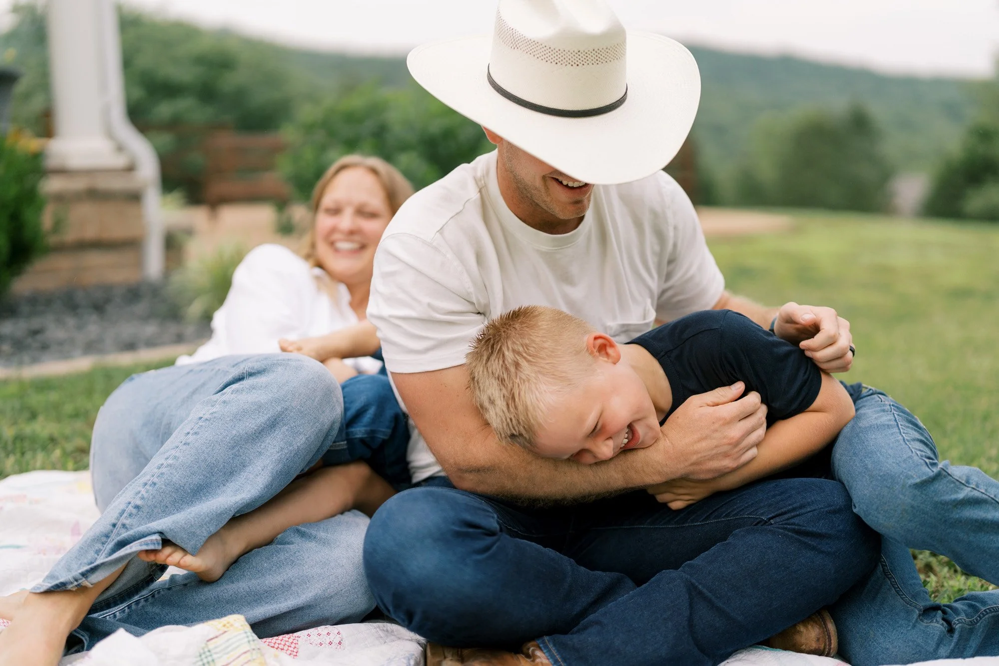 outdoor portrait of a mom and dad sitting on the ground with two kids playing and wrestling on them while smiling and laughing