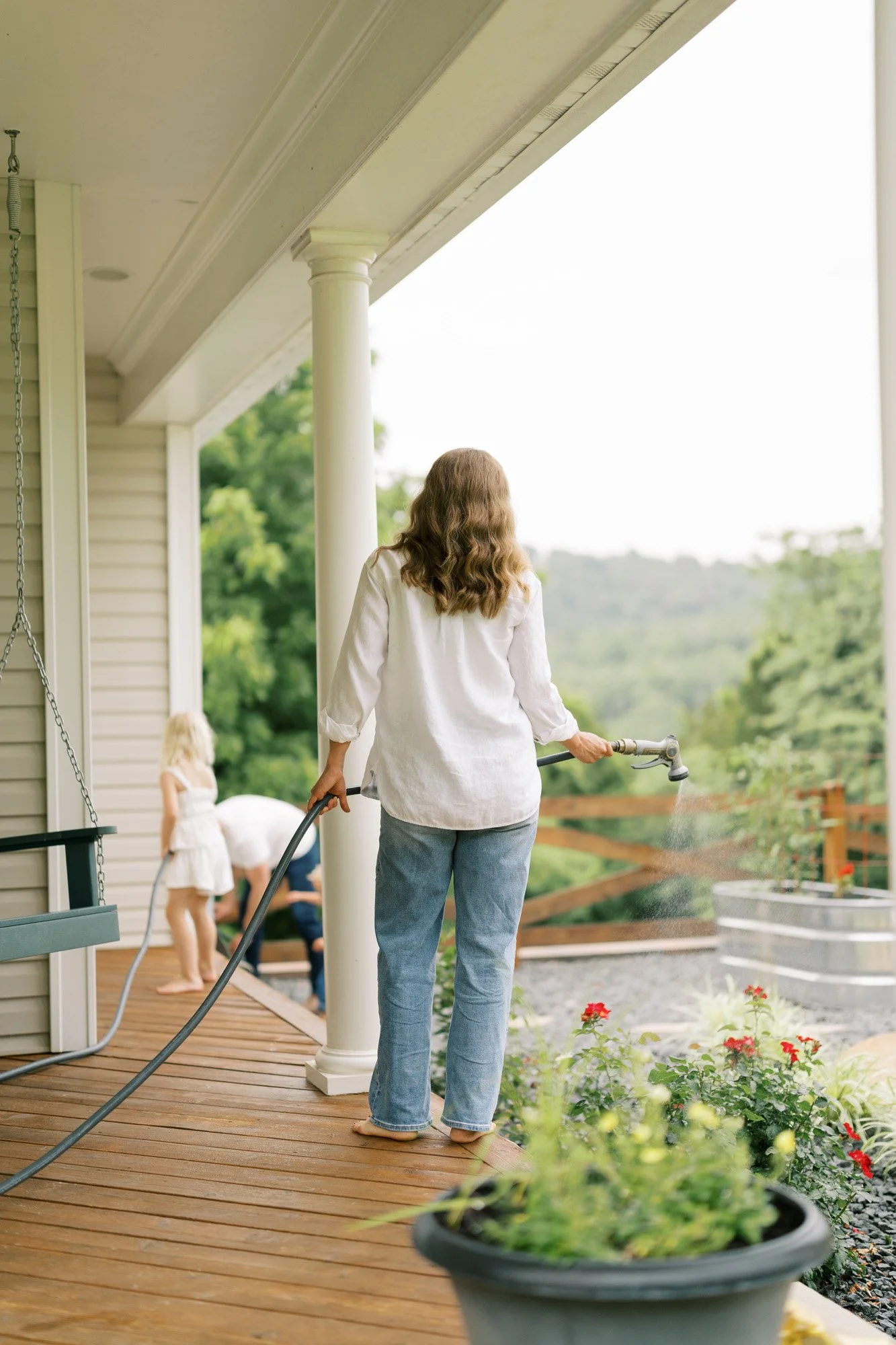 Mom in white shirt and jeans watering plants from a hose with kids helping in the background