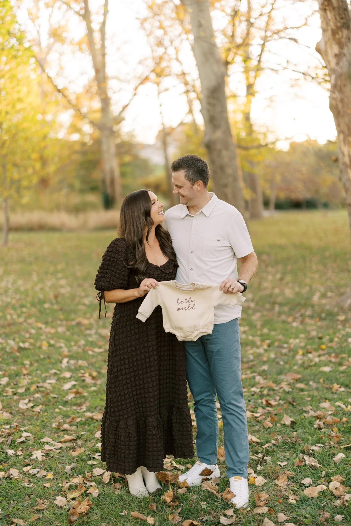 Man and woman standing in a park looking at each other and holding a baby onesie that announces their pregnancy with "hello world"
