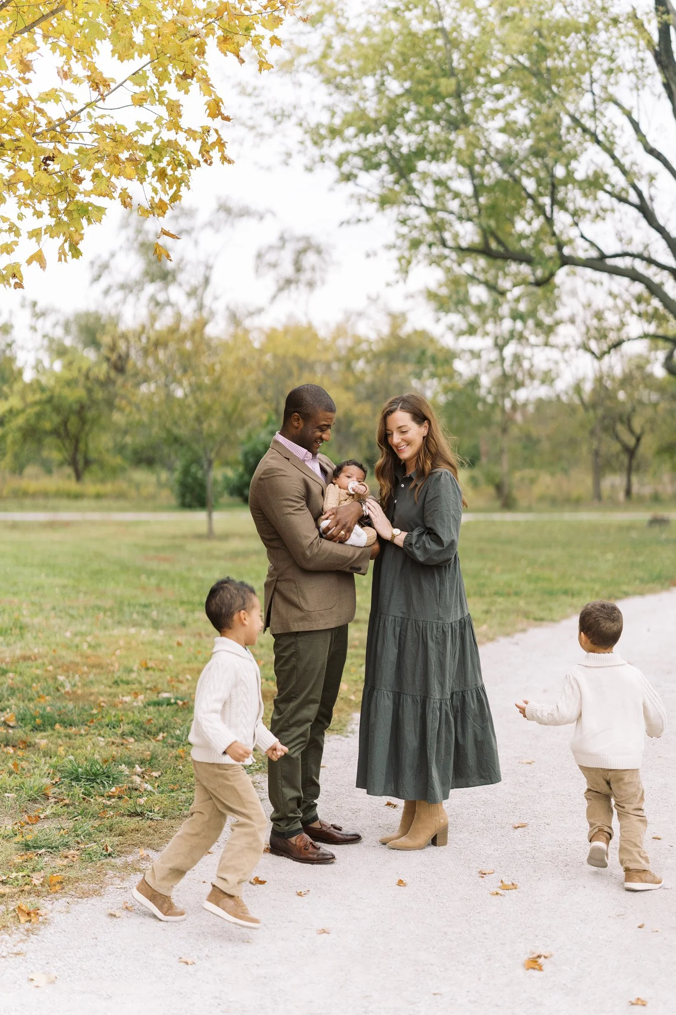 Two parents holding an infant on a path in a park while two toddler sons run around them smiling