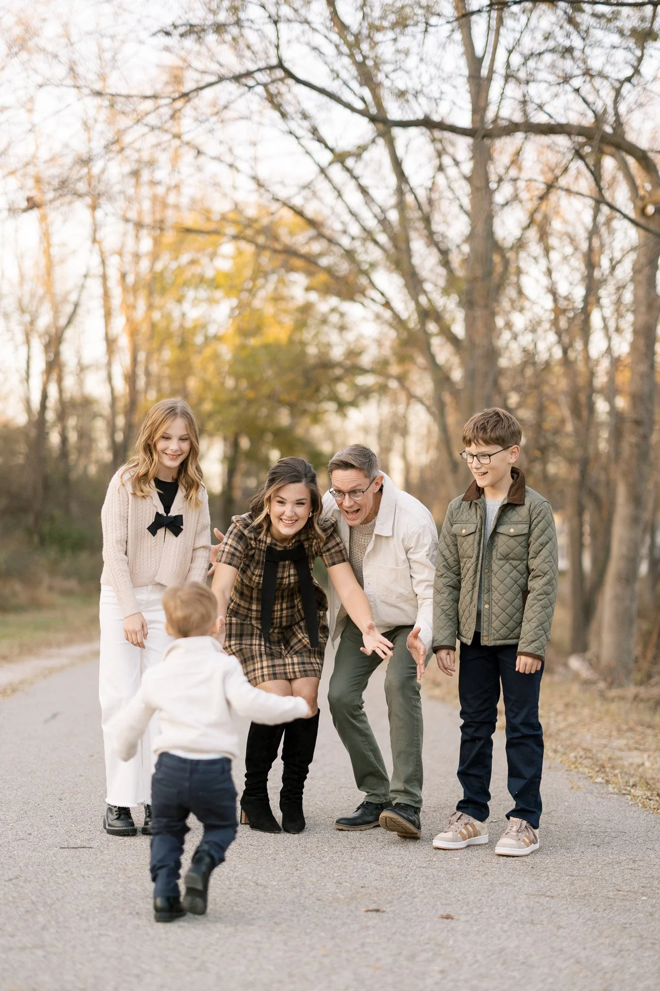 Family of 5 standing on a path in a park in beautiful fall light with littlest kid running toward standing family