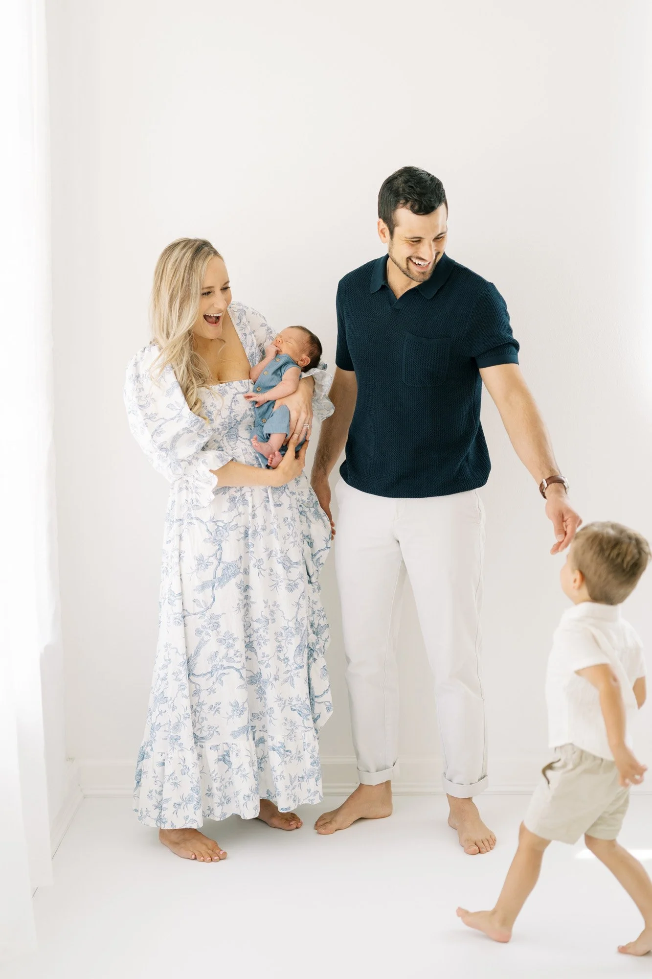 Mom smiling holding newborn son, dad standing next to her holding big brother's hand and smiling