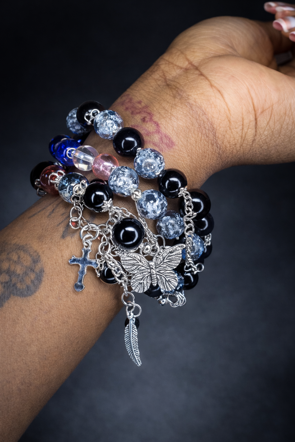 Multiple layered bracelets with assorted beads, charms including a cross, butterfly, and feather, worn on a person's wrist with a tattoo on their arm.