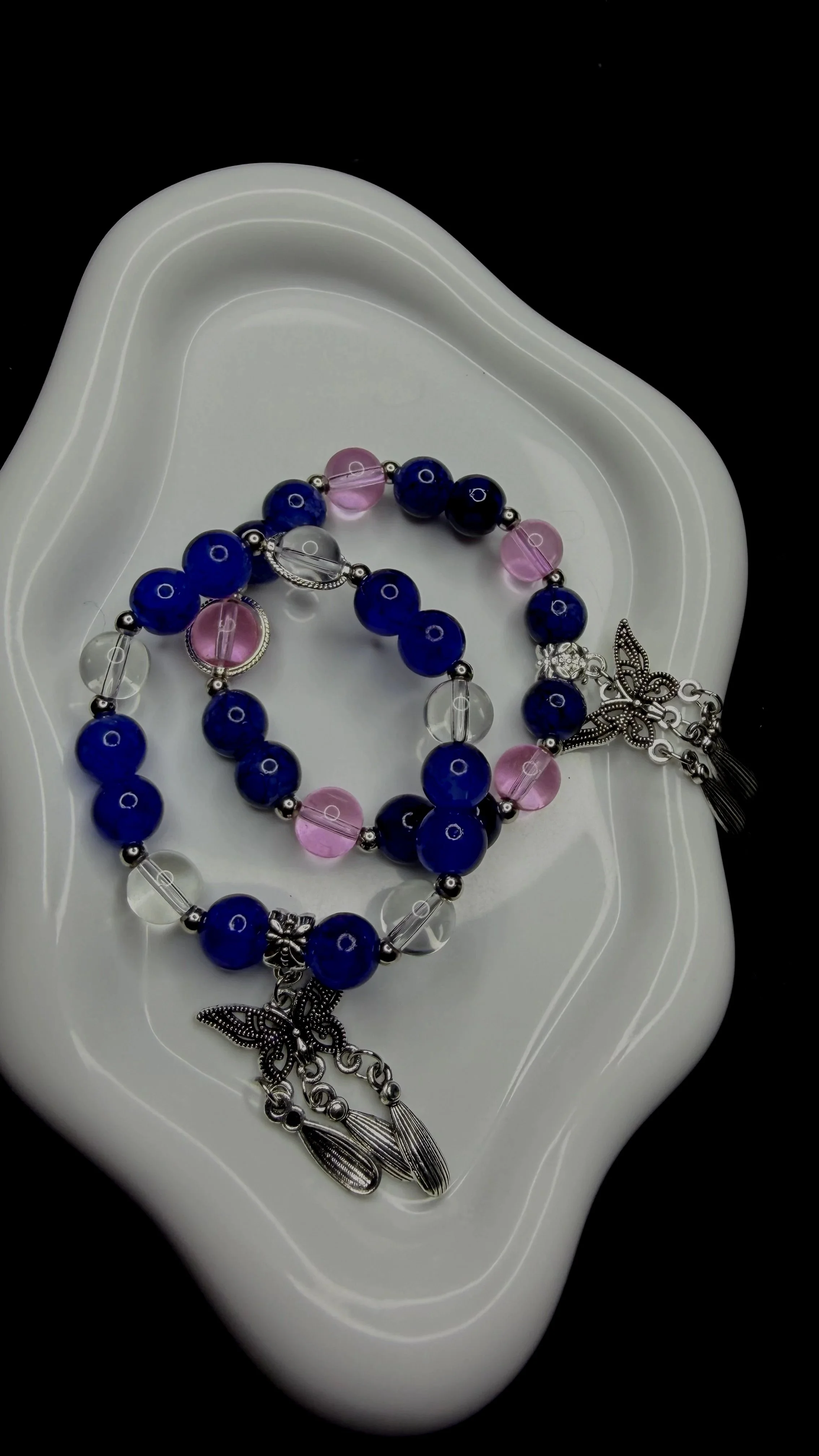A beaded bracelet with blue, pink, and clear beads, decorated with a silver butterfly charm and other silver charms, resting on a white dish against a black background.