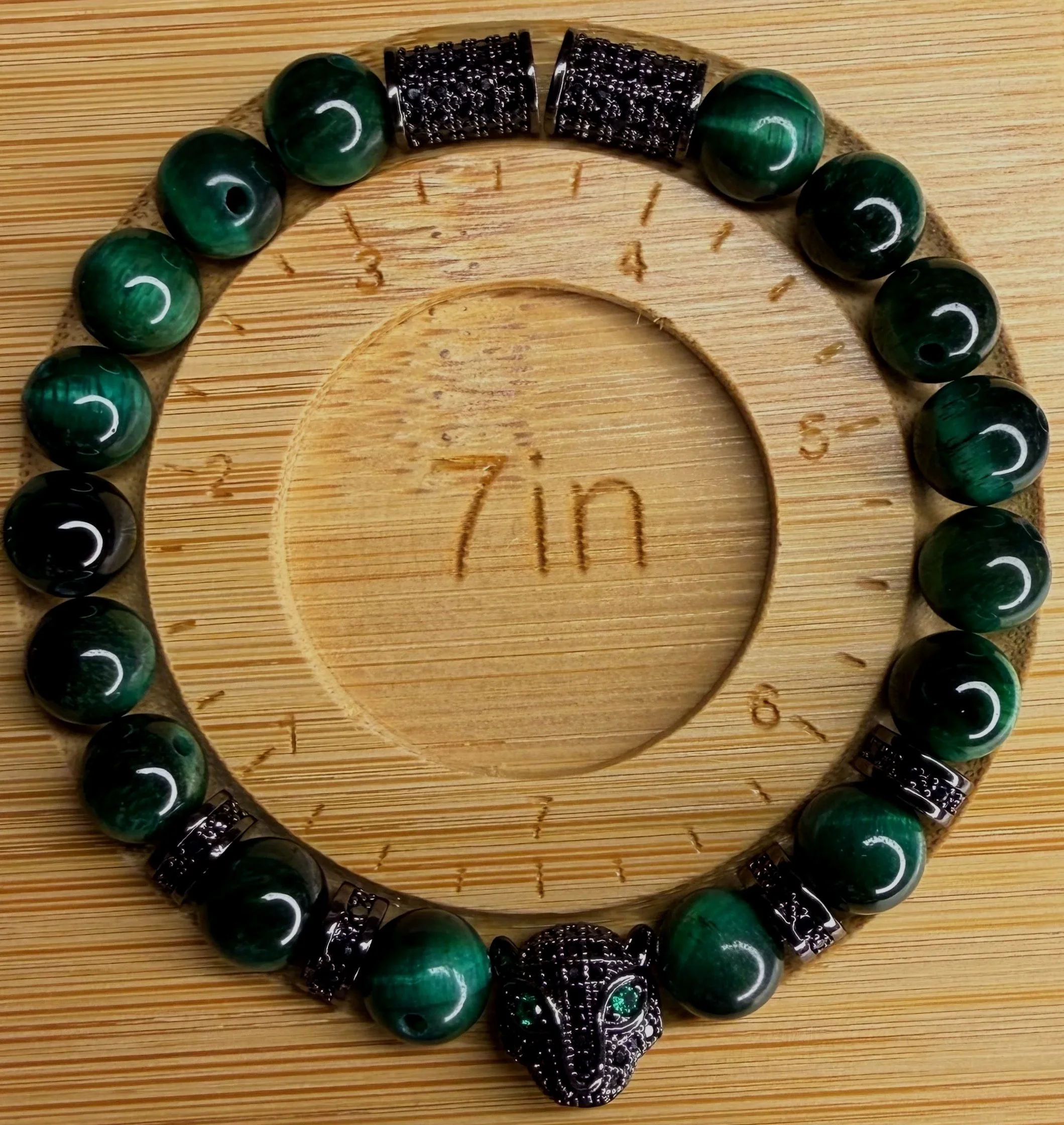 A beaded bracelet with green and black beads and a decorative black bead centerpiece, placed on a wooden surface with the word '7in' engraved.