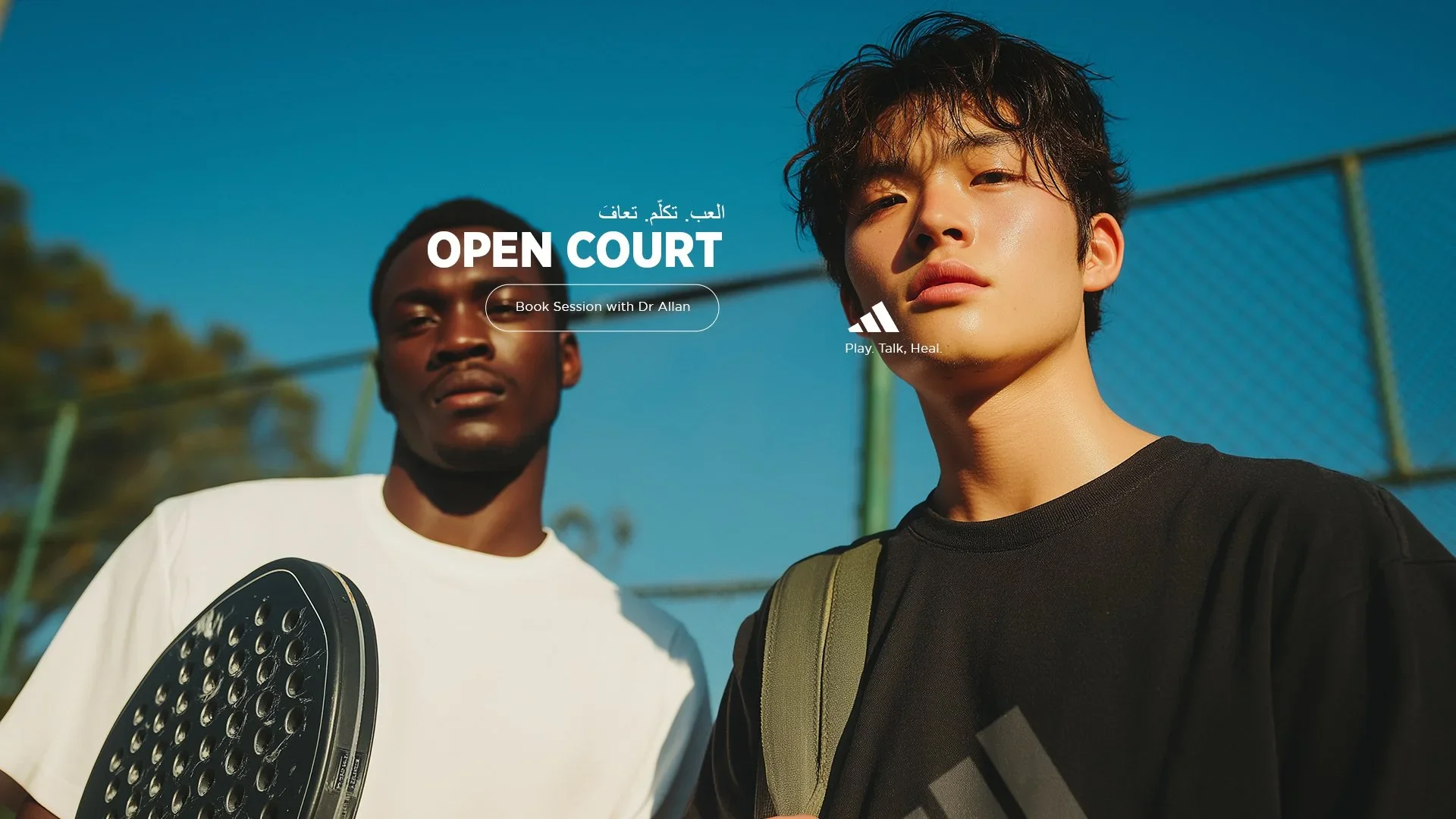 Two young men standing on a tennis court, one holding a tennis racket, with blue sky and a chain-link fence in the background.