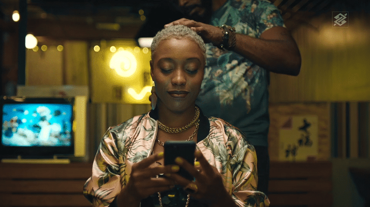 A woman with short blonde hair sitting in a dimly lit room, looking at her phone while a man behind her touches her head.