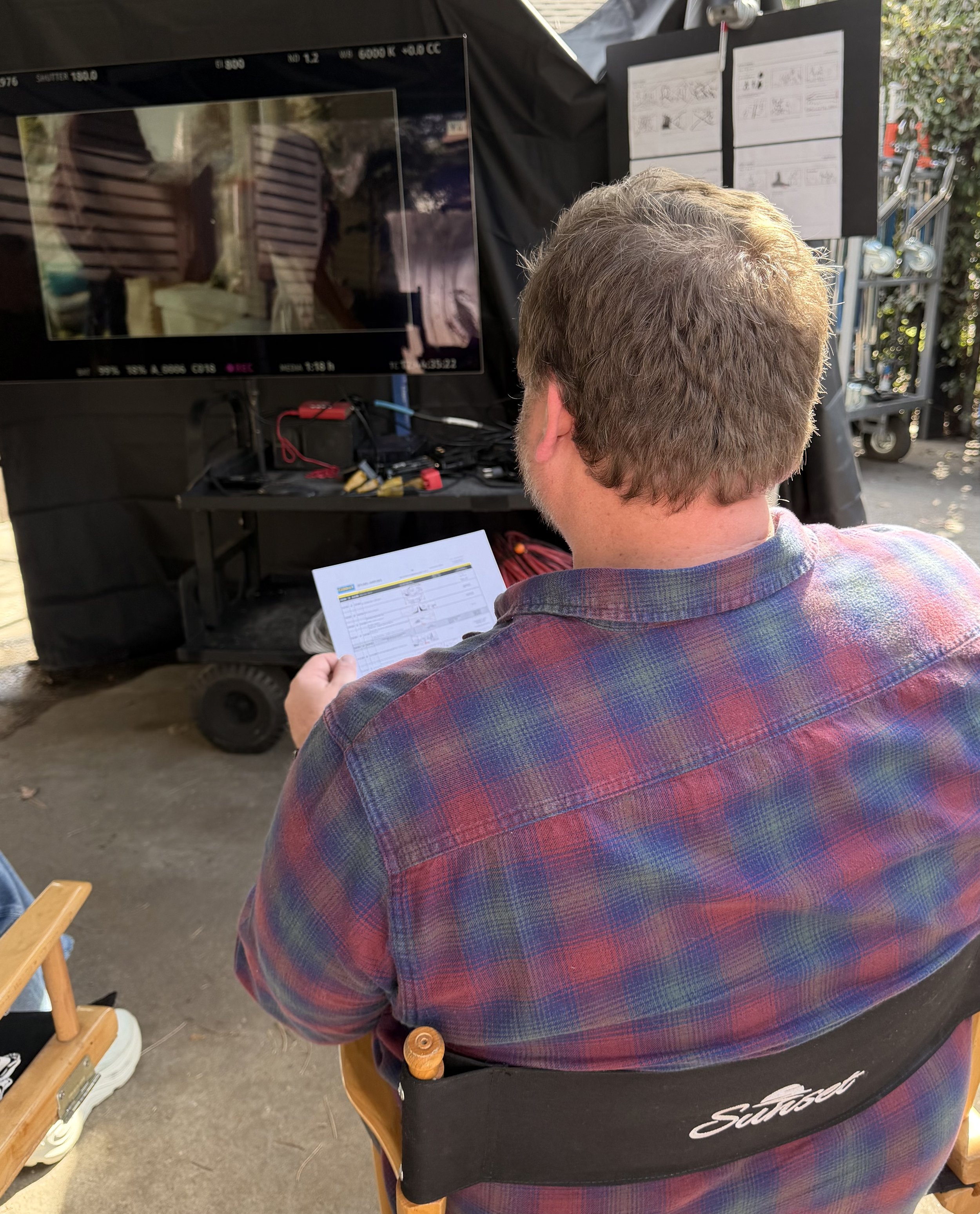 Ted Herteg sitting in a director's chair, looking at a clapperboard and holding a sheet of paper, with a monitor and instructional papers on a black backdrop in an outdoor setting.