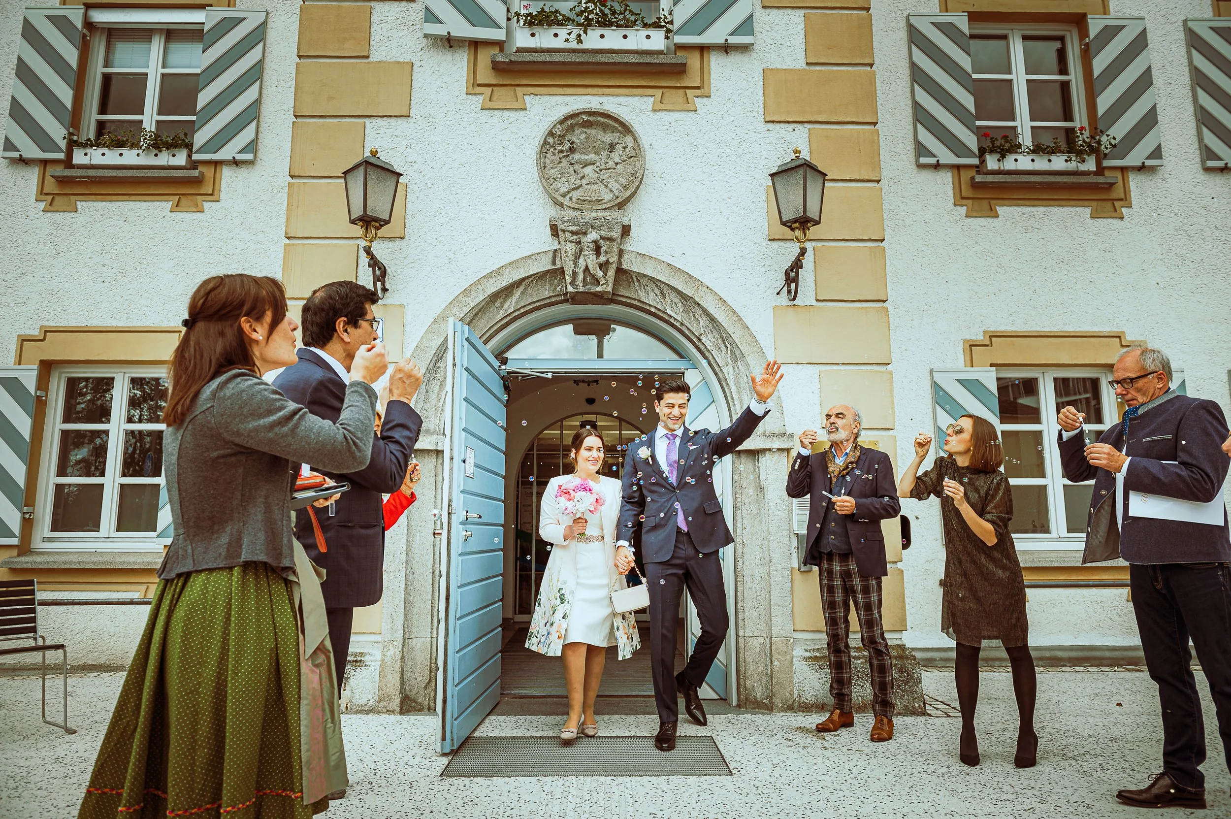A bride and groom are exiting a building through a blue door, celebrating their wedding. The groom, dressed in a suit, is waving and smiling, while the bride, holding a bouquet, follows behind him. Around them, friends and family are cheering, blowing bubbles, and celebrating outside the building.