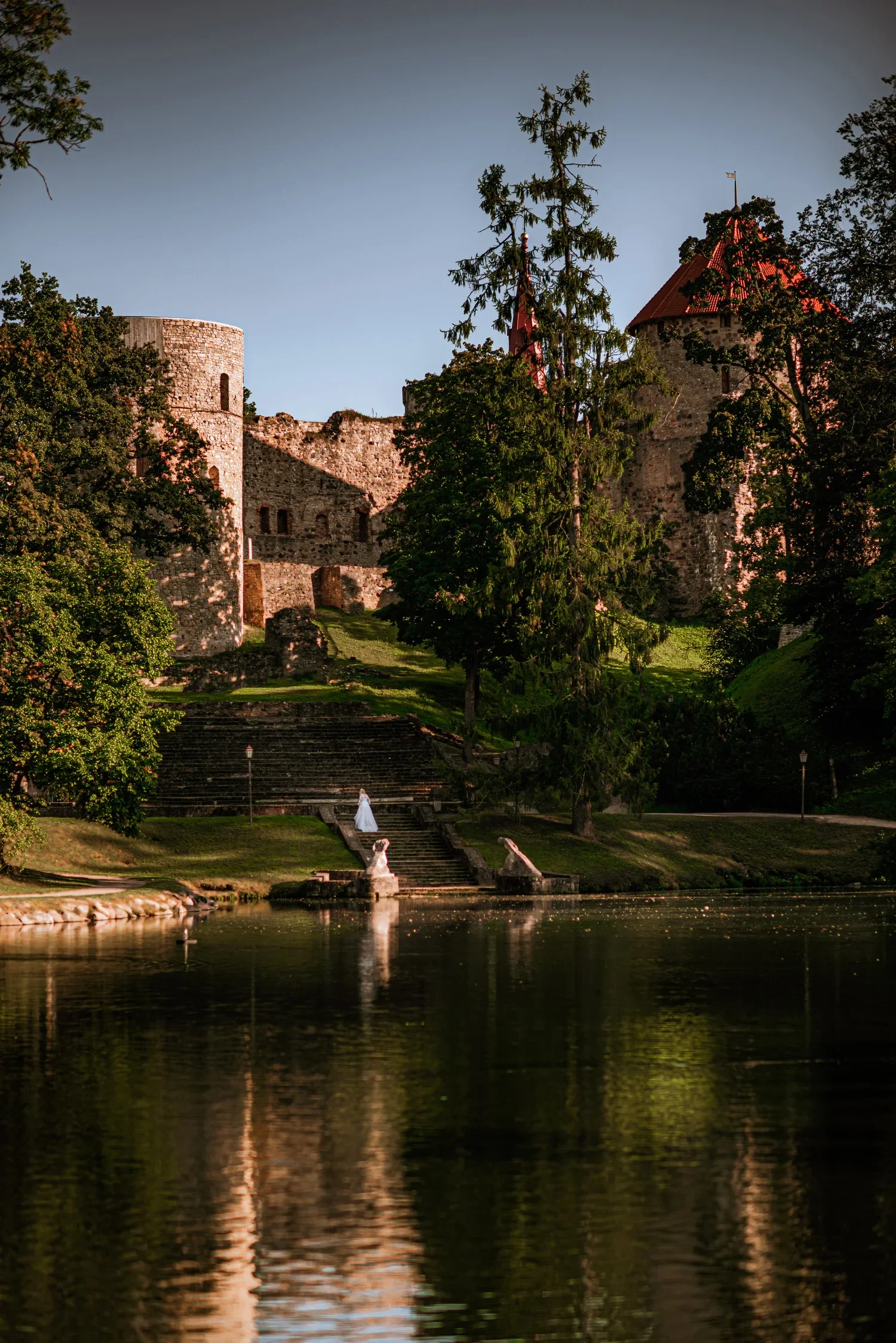 A historic stone castle with towers and turrets on a hill, surrounded by trees, overlooking a reflective lake with stone stairs leading down to the water, where a bride in a white wedding gown stands at the top of the stairs.