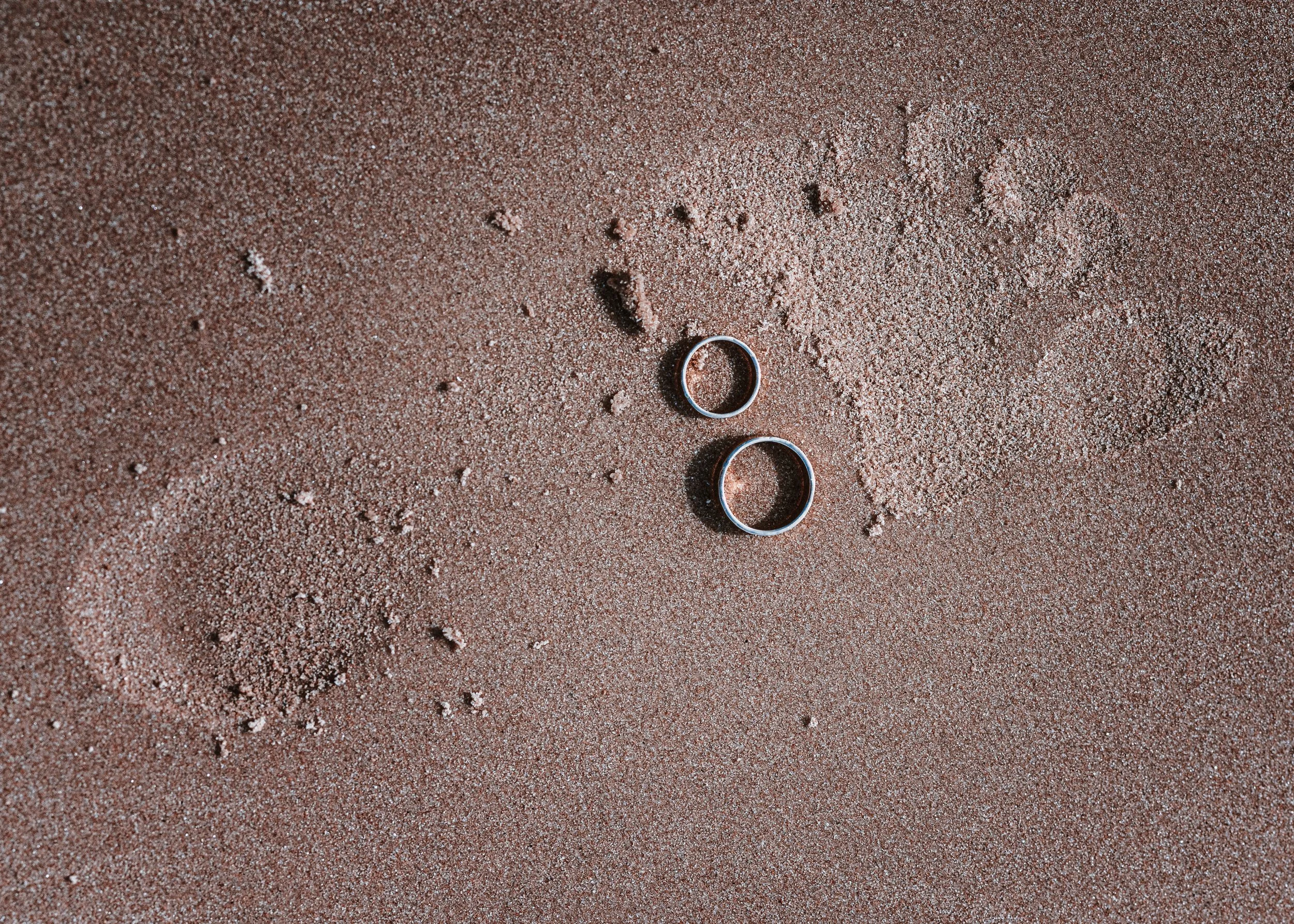 Two wedding rings placed on sandy ground with footprints nearby.
