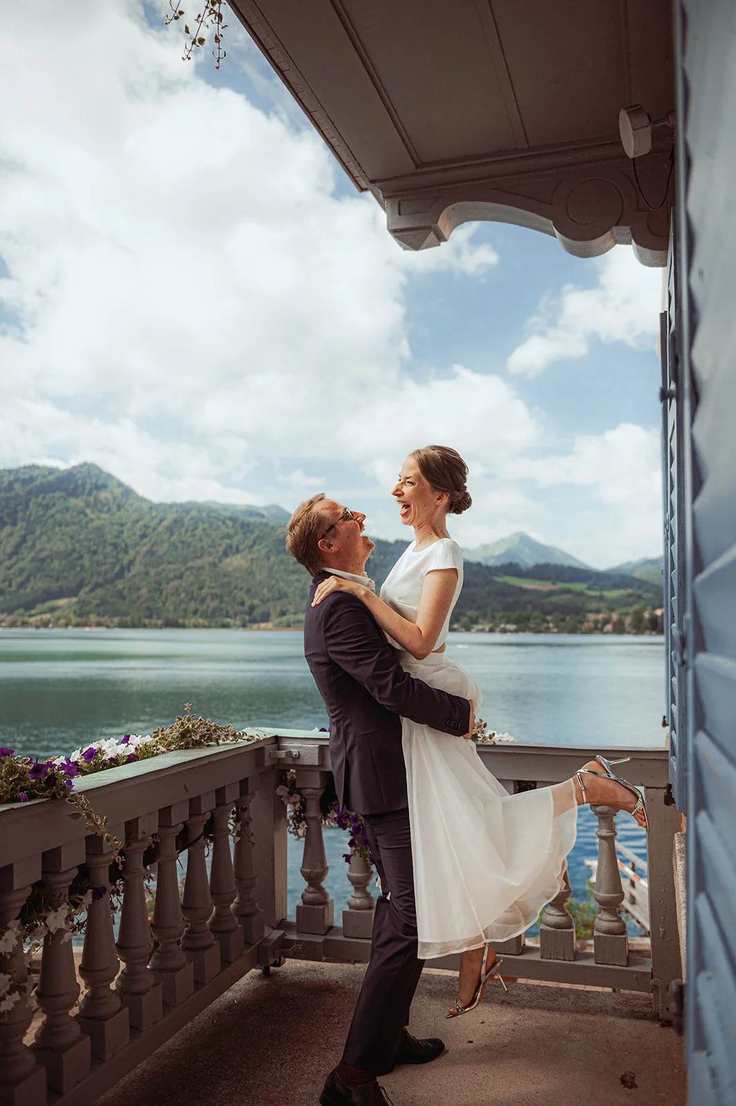 A wedding couple on a balcony overlooking a lake with mountains in the background, with the groom lifting the bride, both smiling and looking at each other.