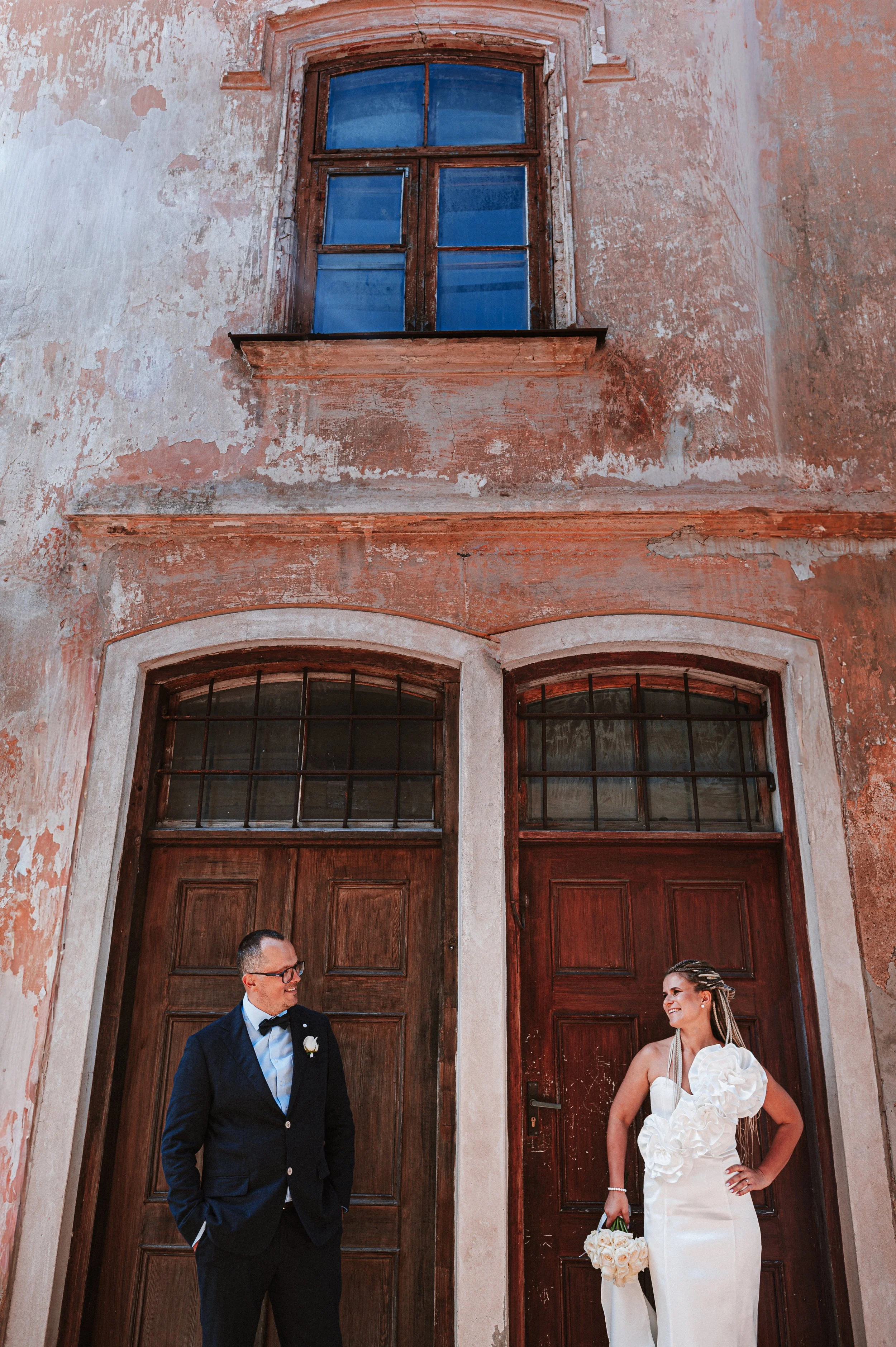 A man and a woman in formal wedding attire standing outside a weathered, rustic building with wooden doors and arched windows, smiling at each other.
