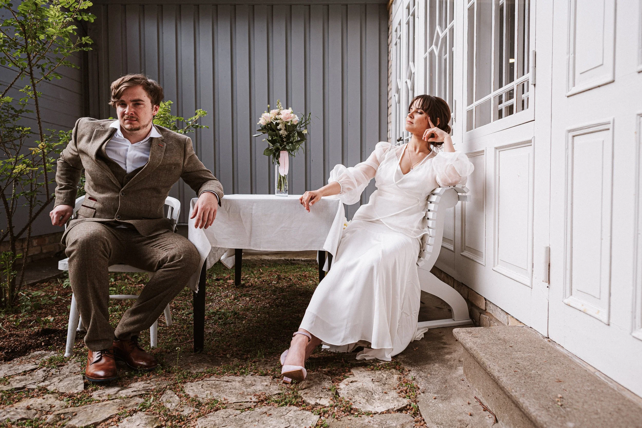 A man in a brown suit and a woman in a white dress sitting outside near a white wooden wall. The woman is reclining and resting her head on her hand. There is a small table with a vase of flowers between them.