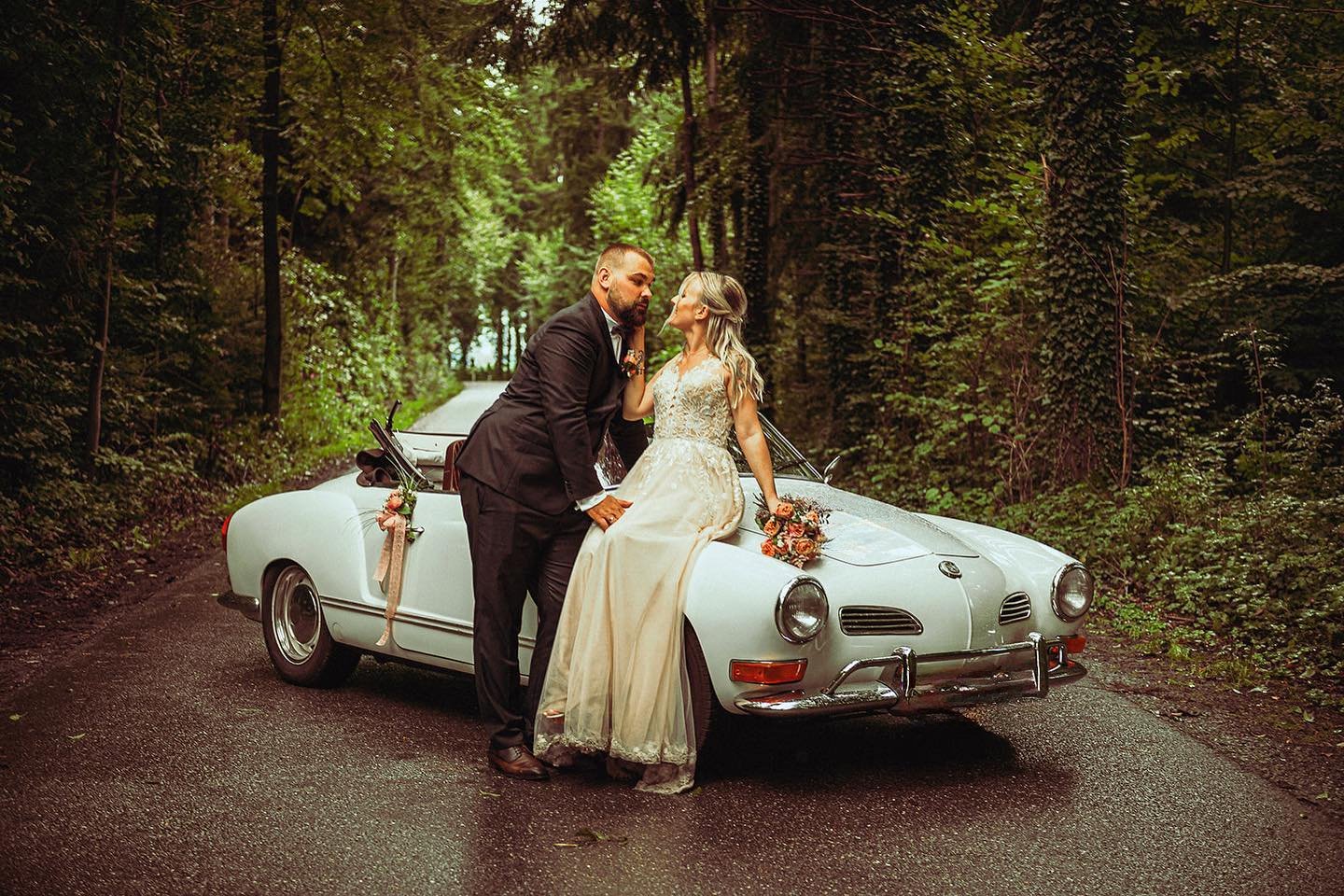 A bride and groom on their wedding day, posing in front of a vintage white convertible car, on a wooded road surrounded by green trees.